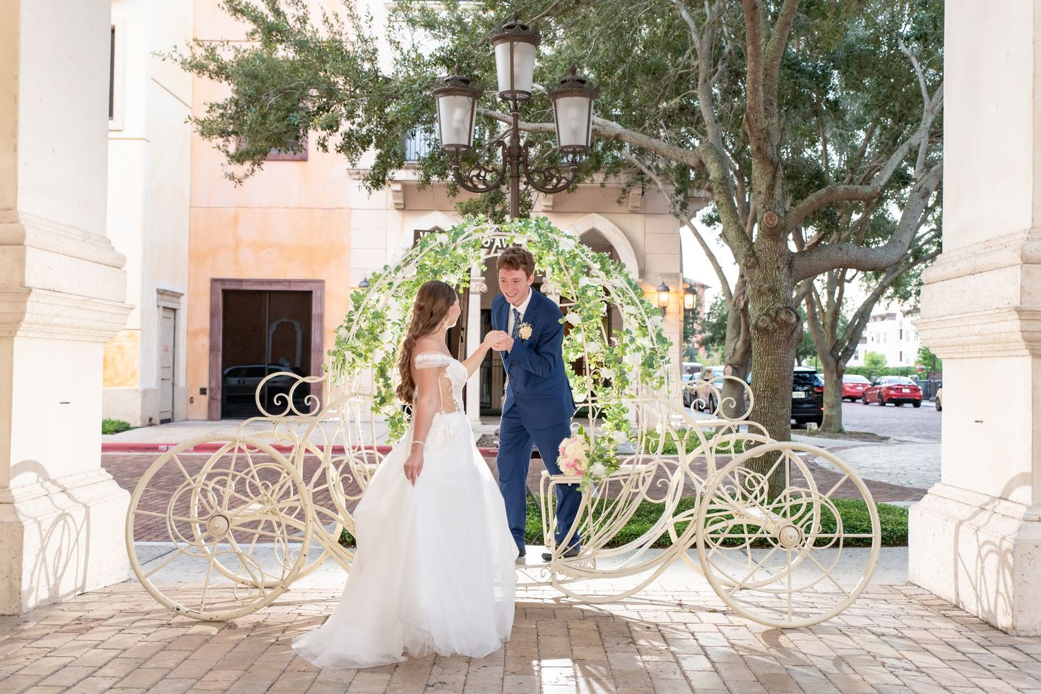 Bride and groom stand beside a white Cinderella carriage outside an elegant venue.