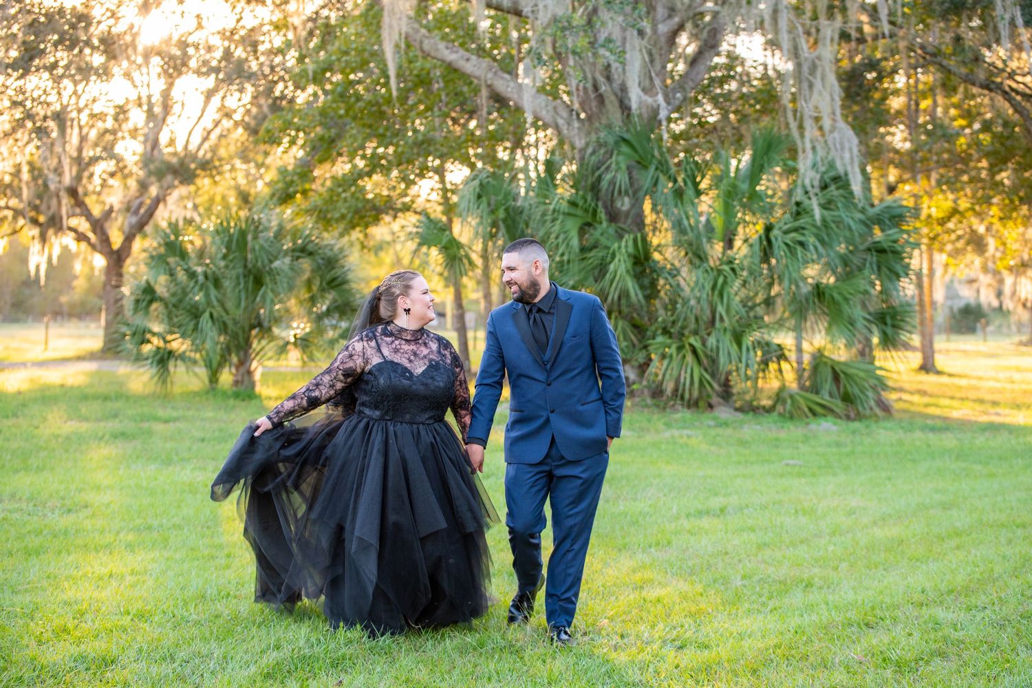 Couple walking hand-in-hand outdoors, woman in black gown and man in blue suit, surrounded by lush green trees.