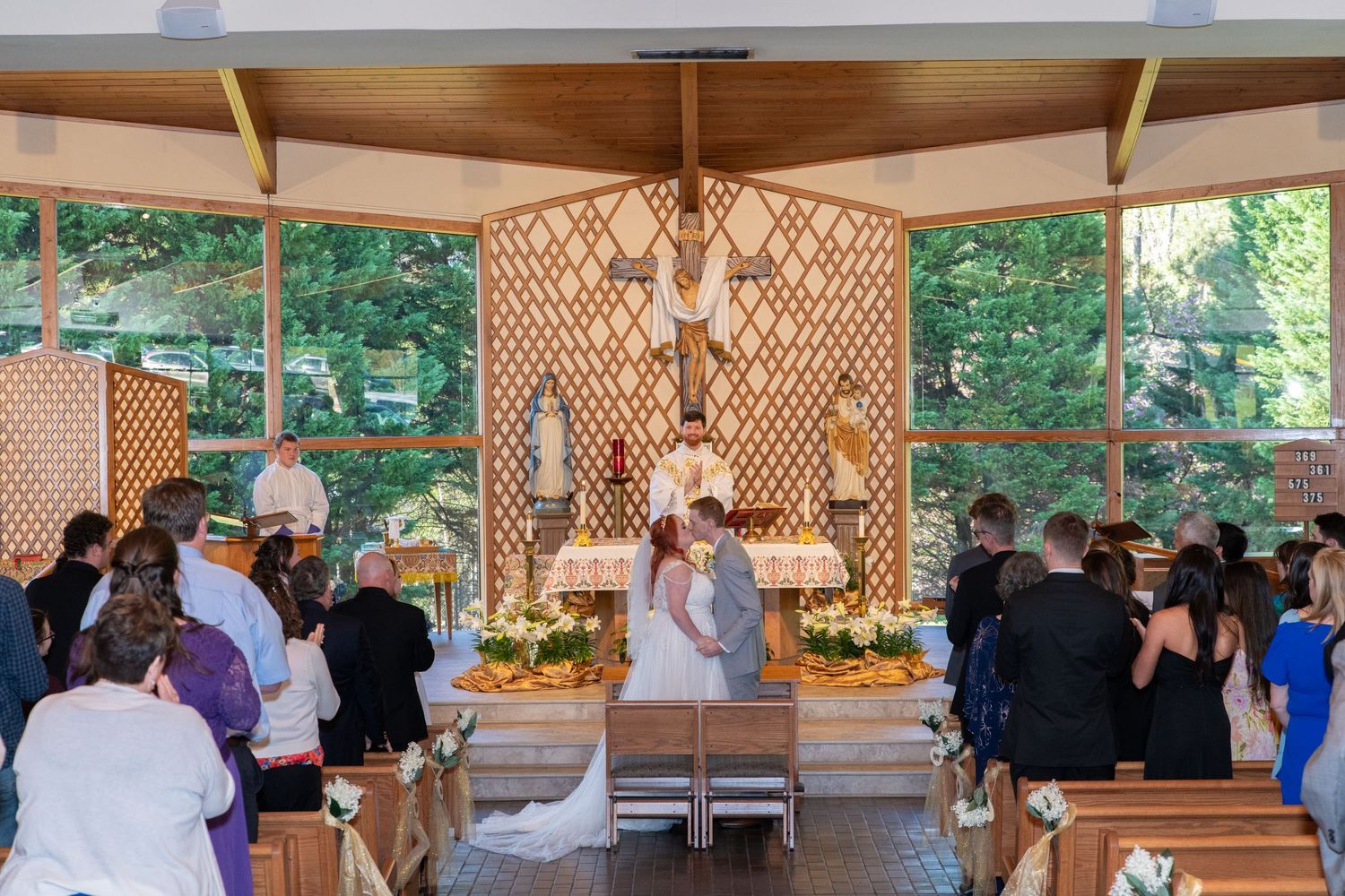A bride and groom stand at the altar during a wedding ceremony in a modern chapel with wooden lattice walls and large windows.