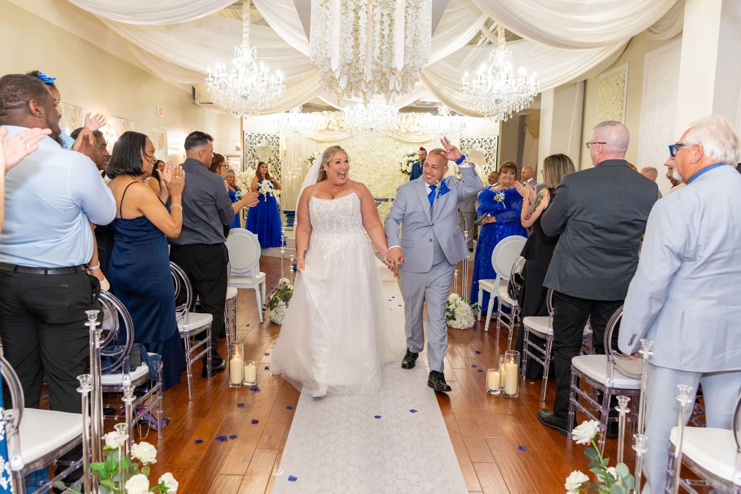 Newlywed couple walks down the aisle at elegant indoor wedding ceremony with guests cheering.