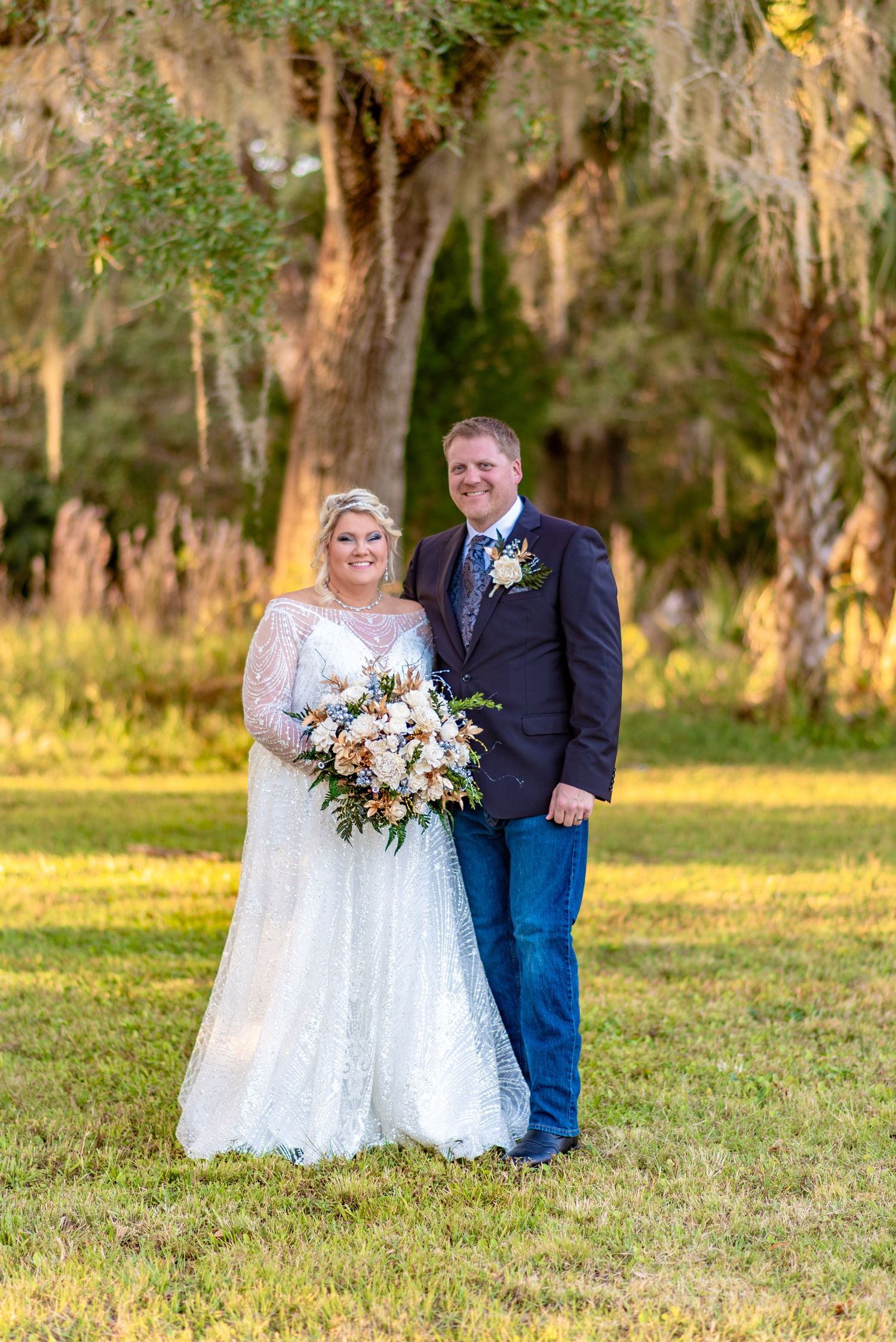 Bride in lace gown and groom in navy suit pose together under Spanish moss trees at sunset.