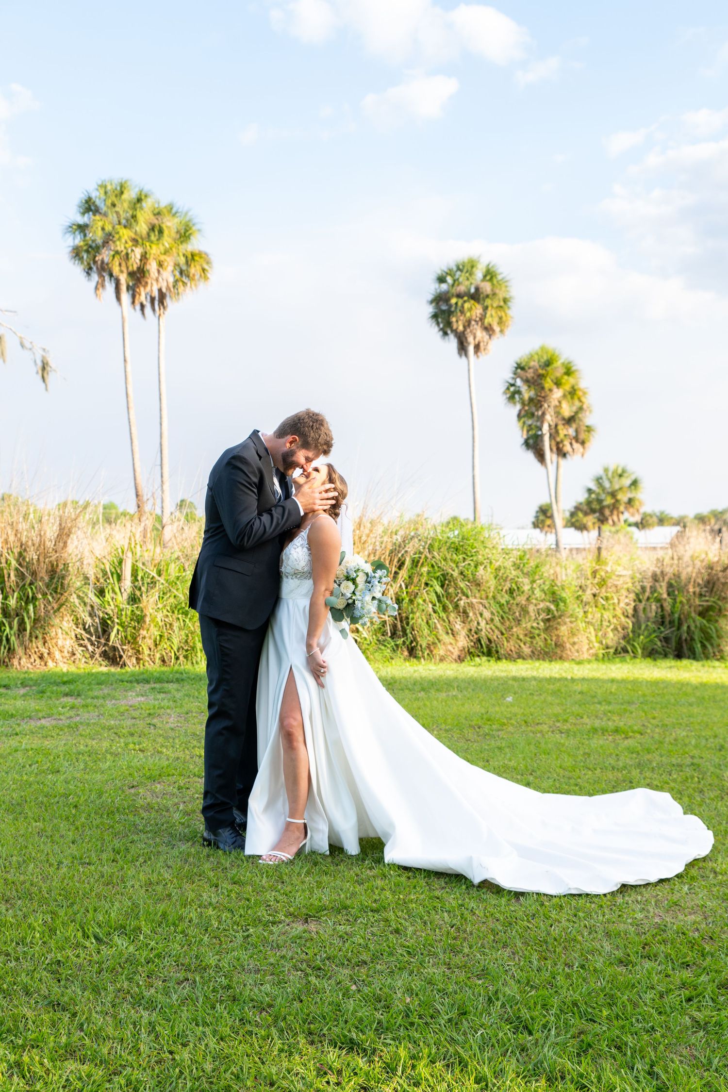 Bride and groom share a romantic kiss on green lawn with tall palm trees and blue sky backdrop at outdoor wedding.