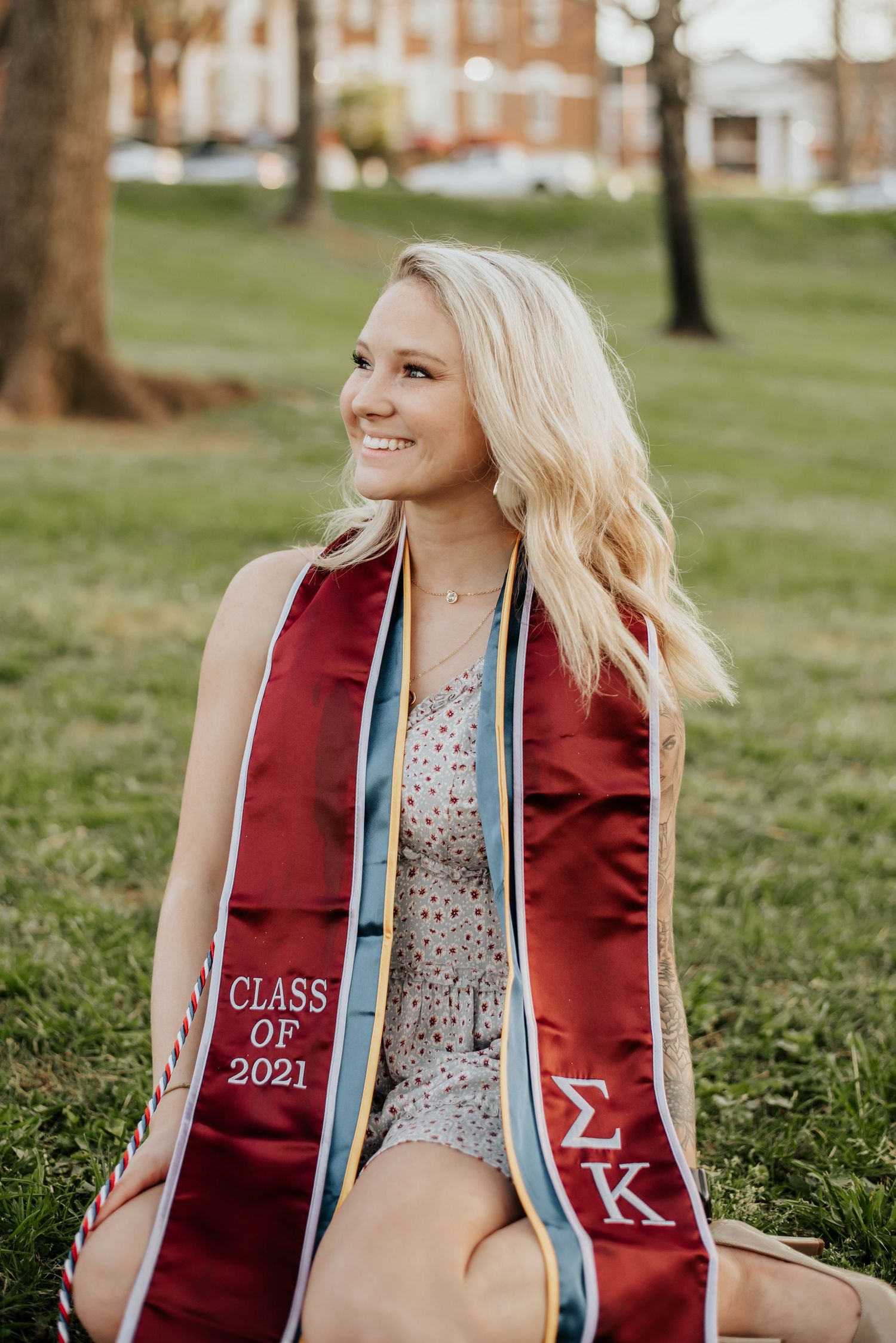 Smiling blonde graduate sitting on grass wearing Sigma Kappa Class of 2021 graduation stole.