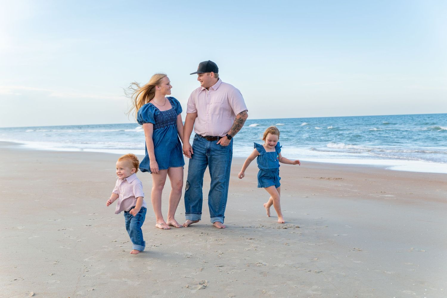 Family of four enjoying a sunny beach walk, with two toddlers running along the shoreline near gentle ocean waves.