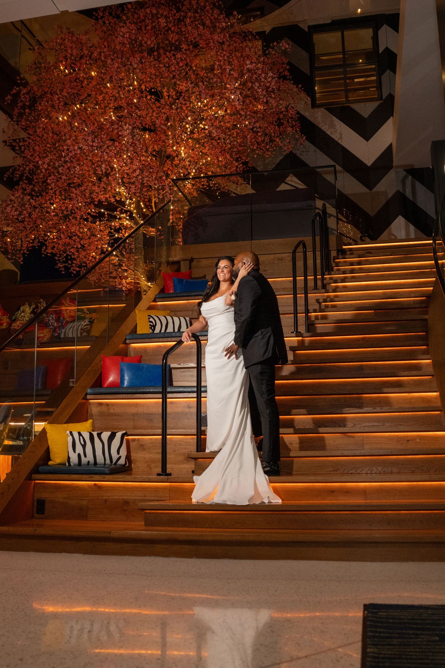 Bride and groom pose on illuminated wooden stairs beside a glowing orange tree in an elegant indoor venue.