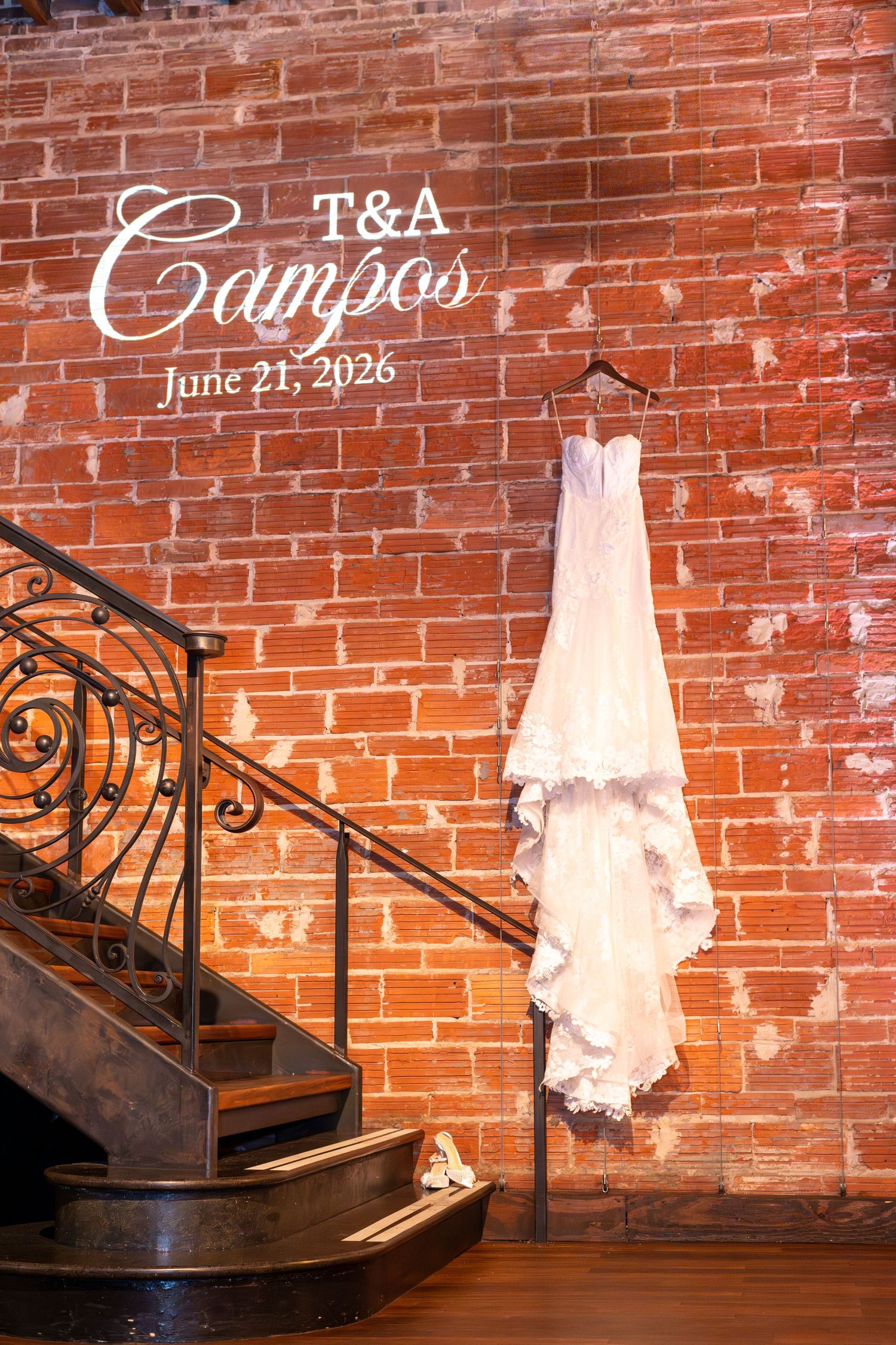 White lace wedding dress hanging on brick wall with T&A Campos June 21 2026 monogram projection near elegant staircase.
