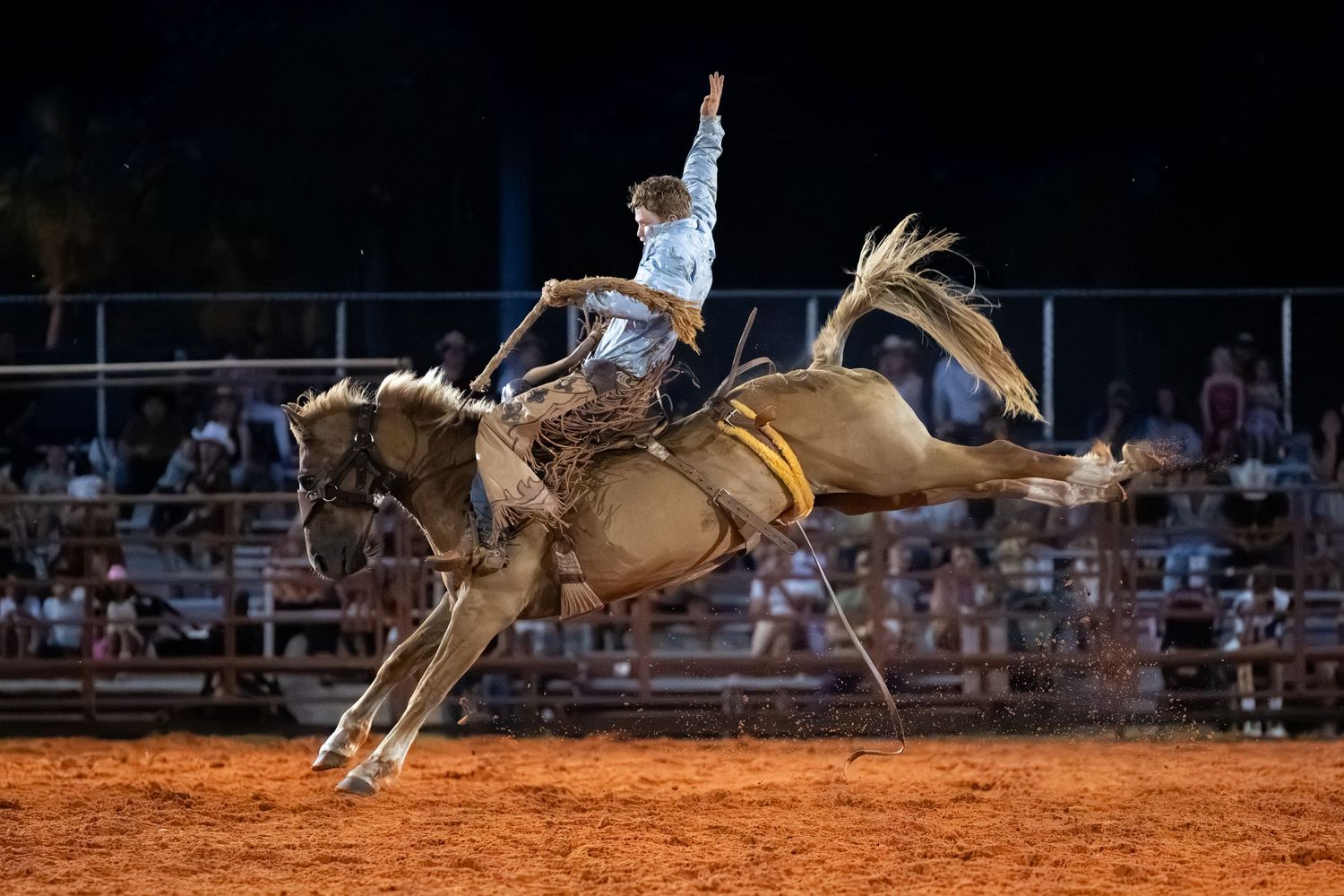 Rodeo cowboy riding a bucking bronco horse at a nighttime rodeo event on a dirt arena.