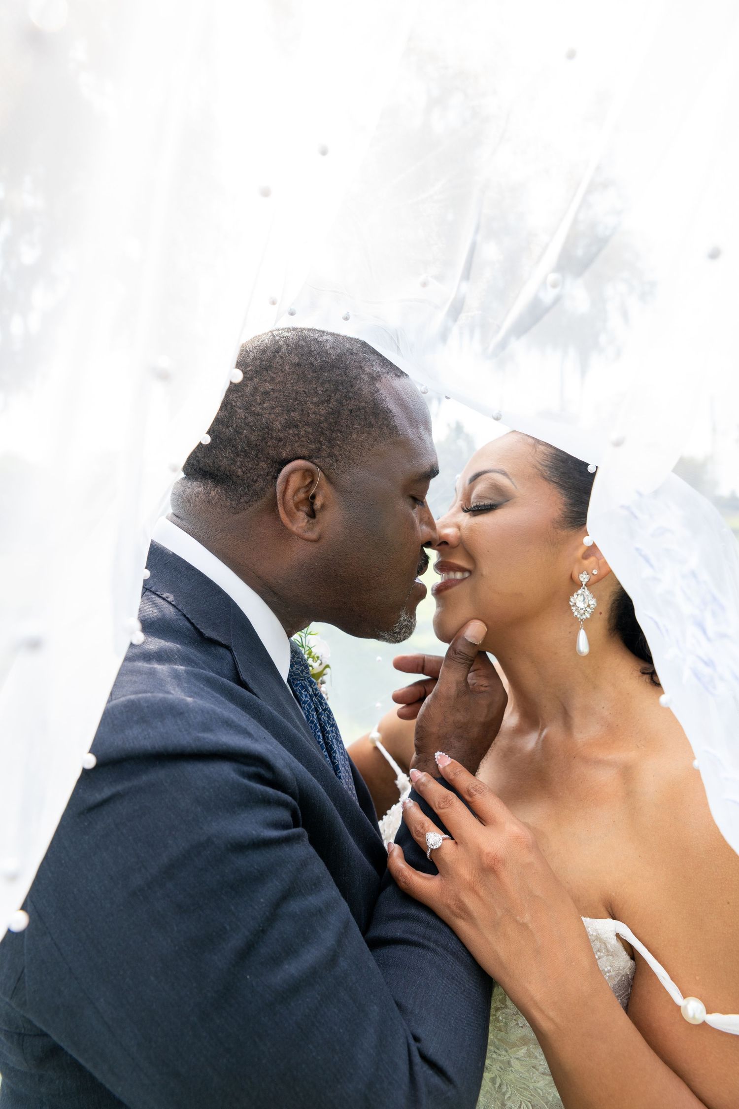 A groom in a navy suit tenderly kisses his bride in a white gown and veil on their wedding day.