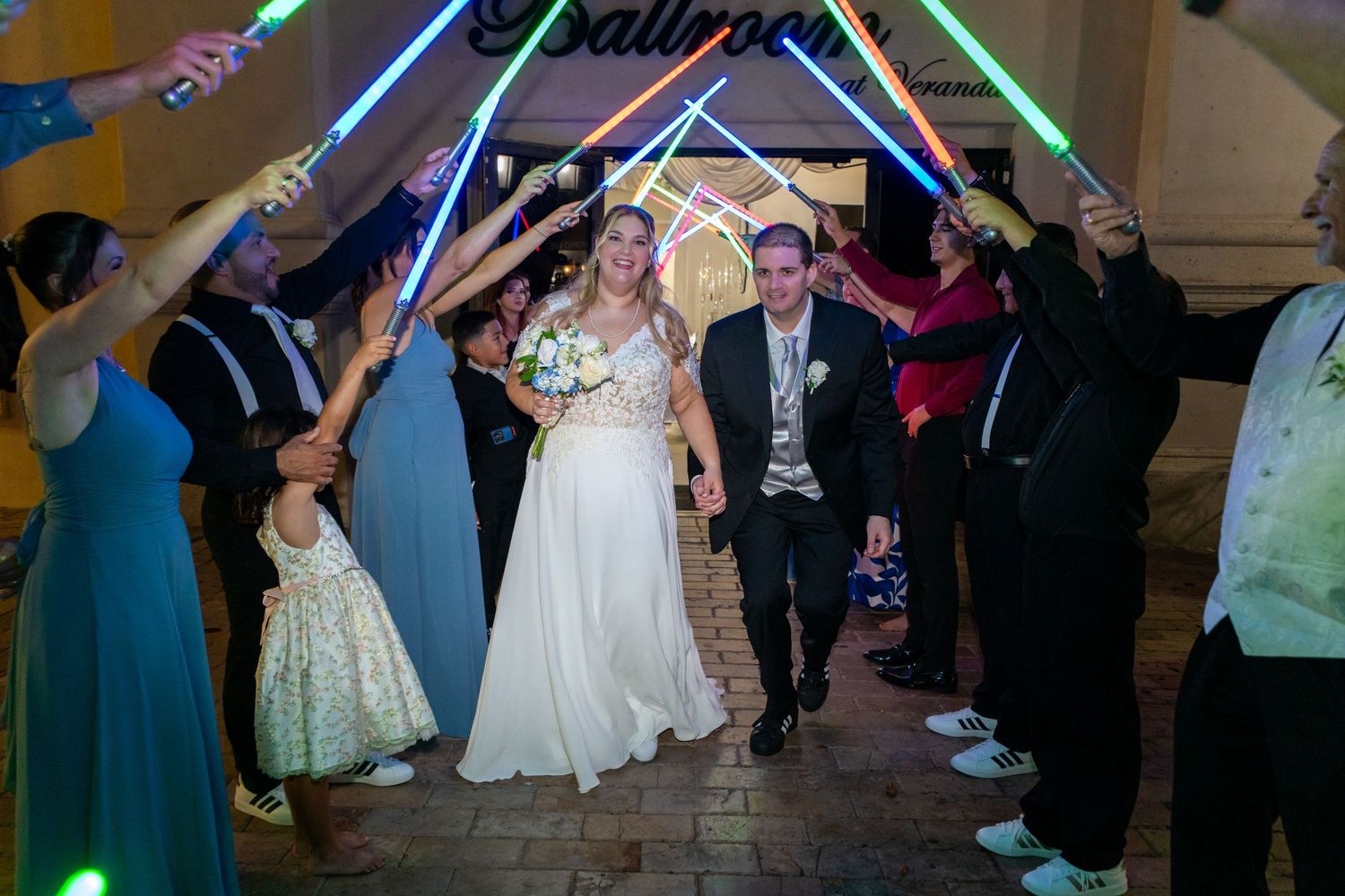 Newlyweds walk through a Star Wars lightsaber arch held by wedding guests at a themed reception.
