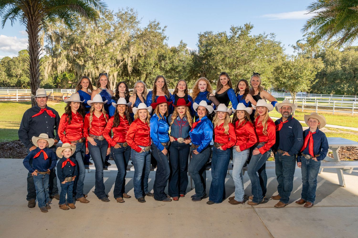 Large group of cowboys and cowgirls in red and blue western outfits posing outdoors at a ranch.