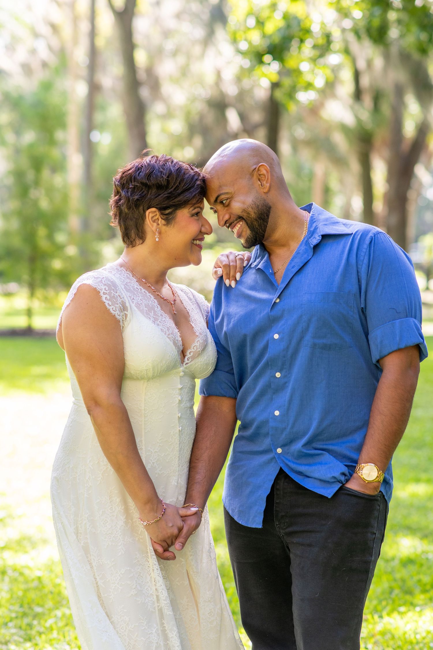 A couple shares a romantic moment outdoors, woman in white lace dress and man in blue shirt, smiling at each other.