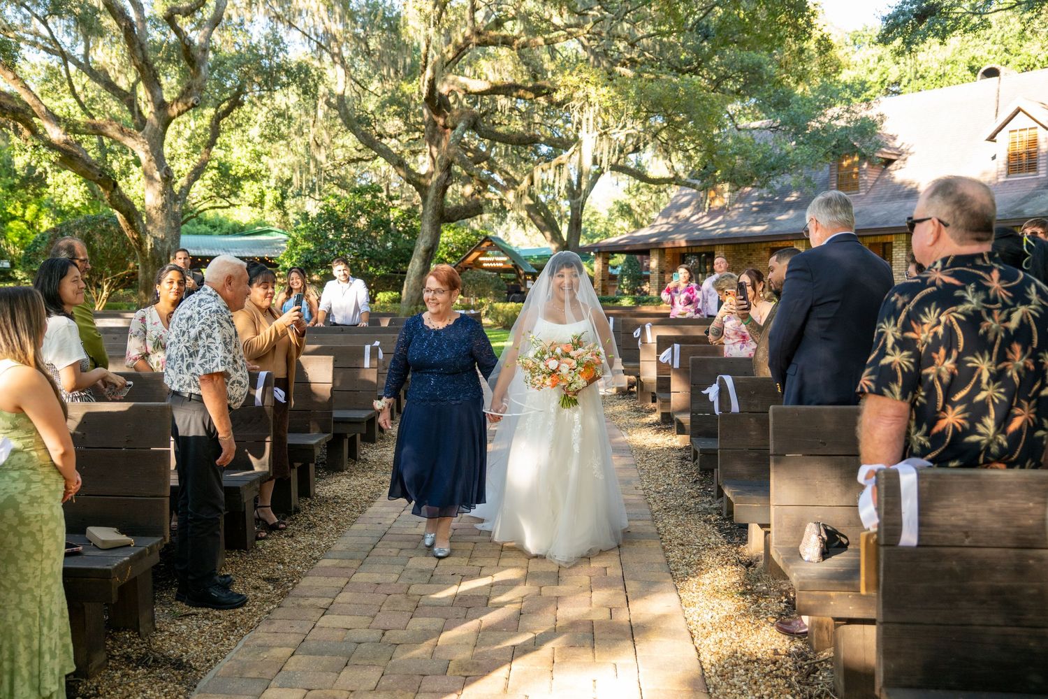 Bride walks down outdoor aisle with escort, surrounded by guests seated on wooden pews under oak trees.