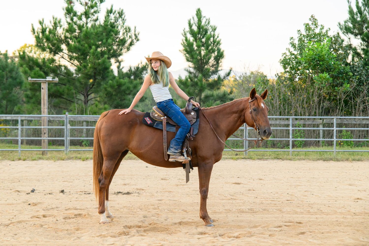 A young woman in a cowboy hat rides a chestnut horse in a sandy arena surrounded by green trees.