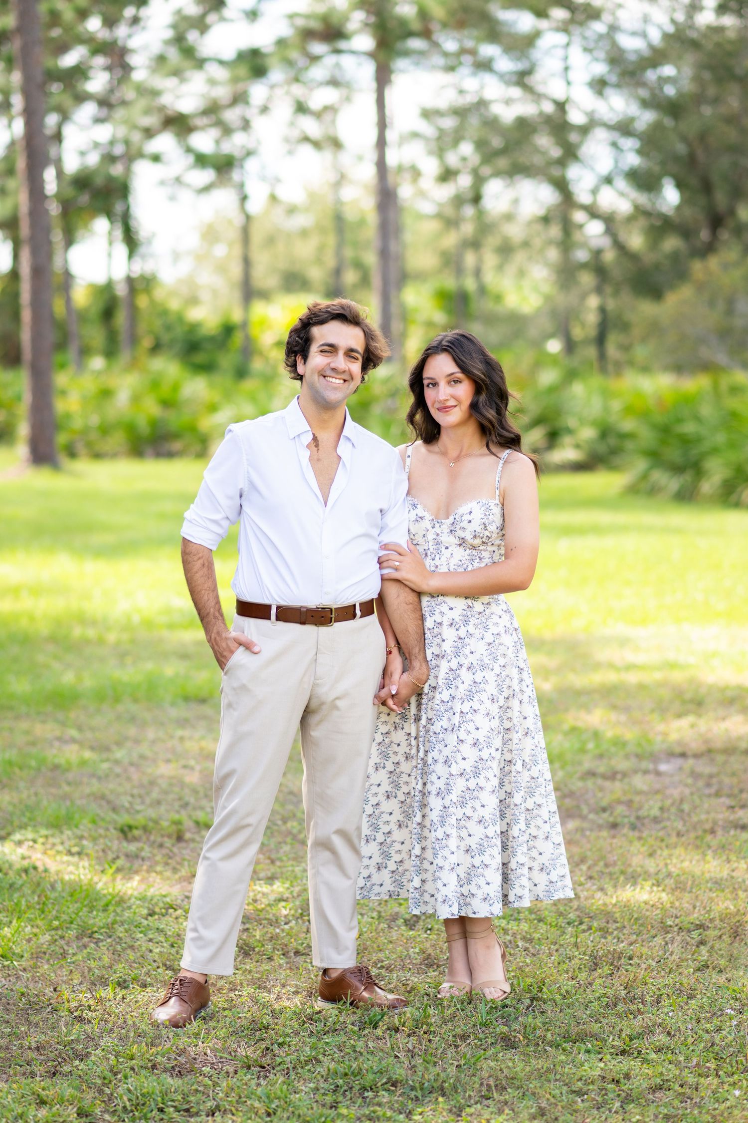 Couple holding hands in a sunny park, man in white shirt and khakis, woman in floral midi dress.