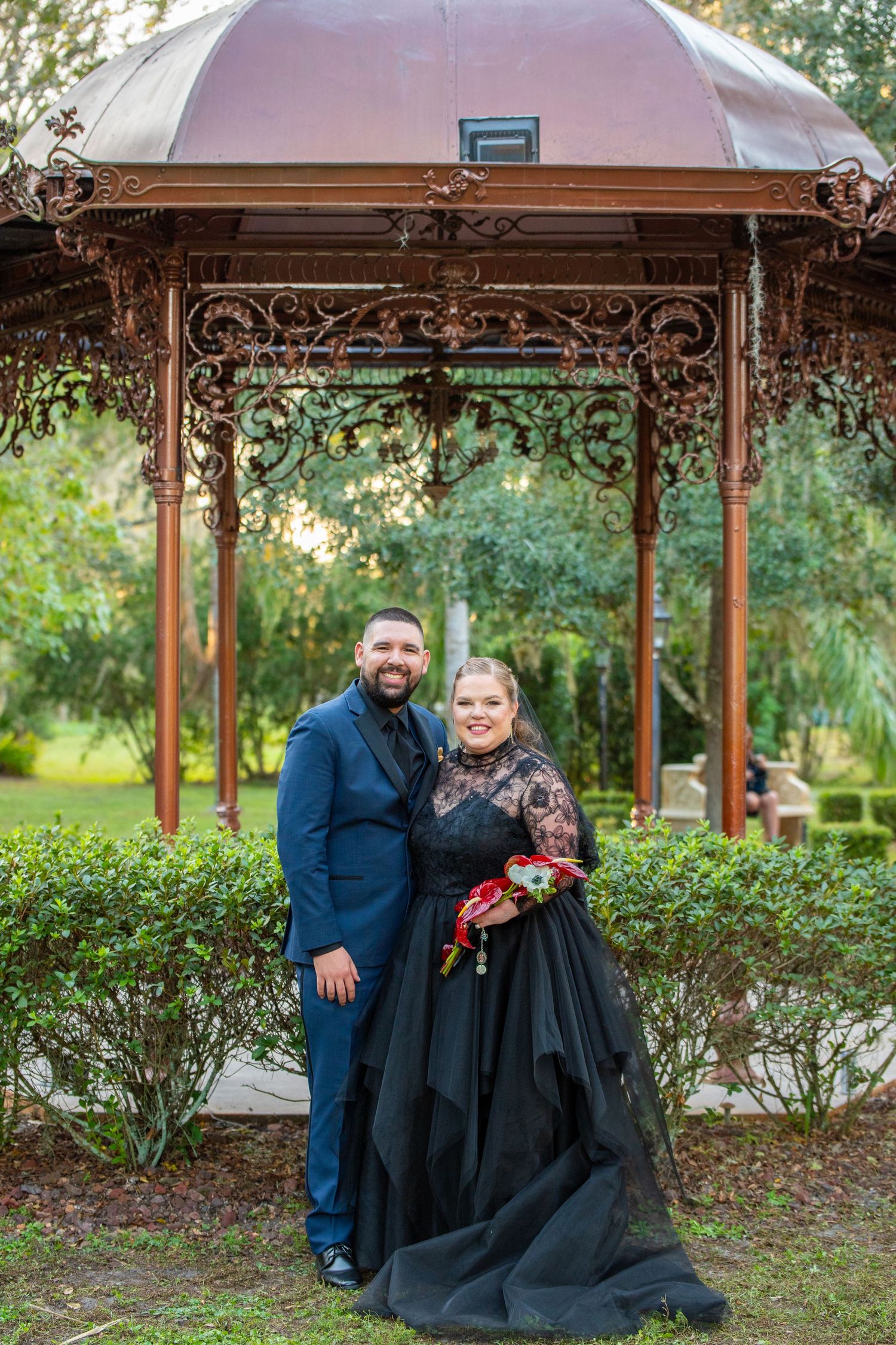 Couple poses in front of ornate gazebo, bride in black lace gown holding bouquet, groom in navy suit.