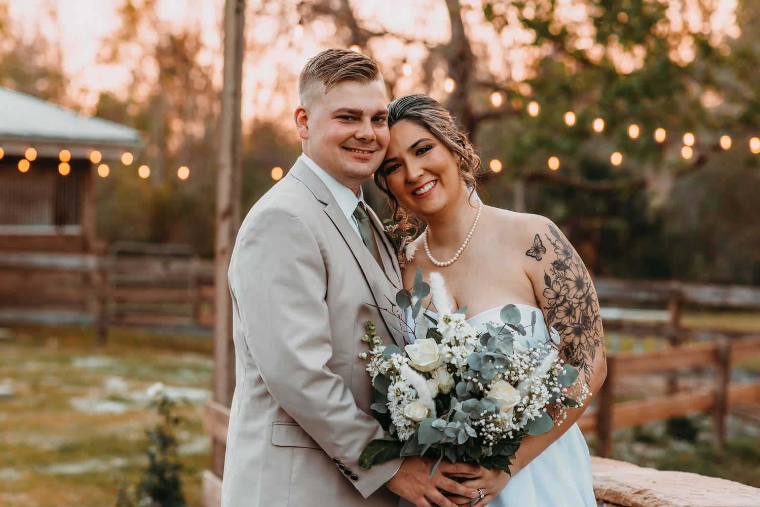 Smiling bride and groom posing outdoors at sunset with white floral bouquet.