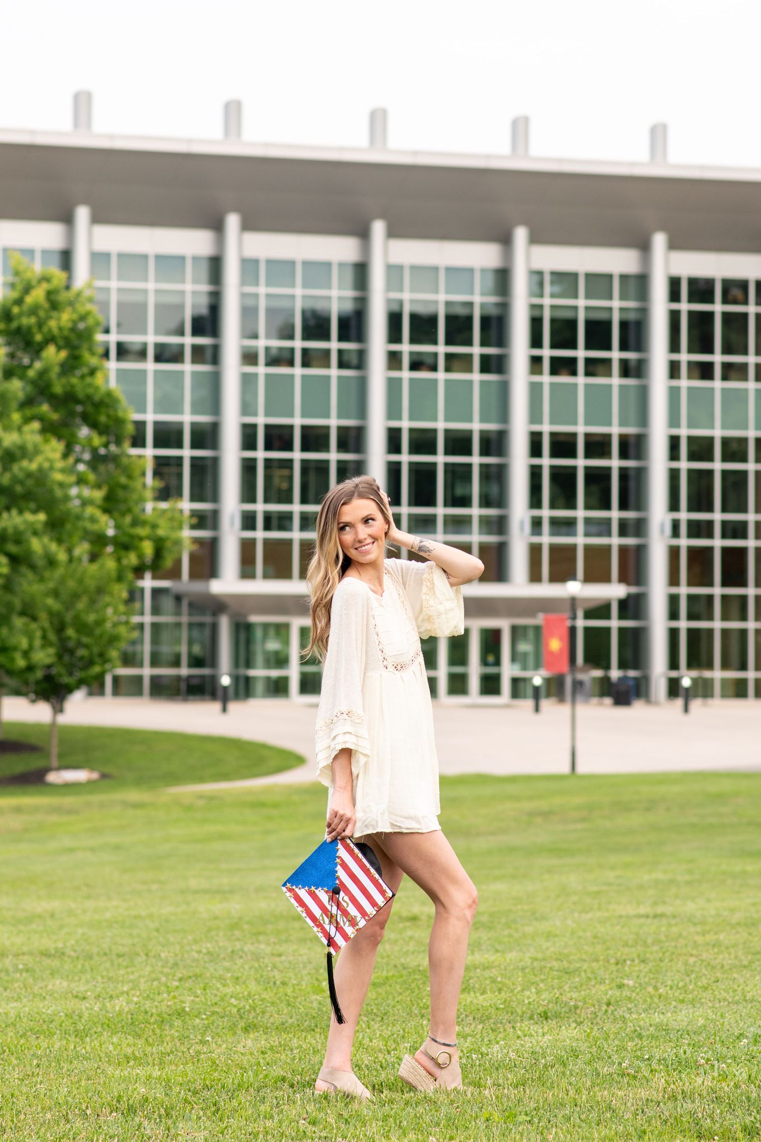 Female graduate in white dress holding American flag posing on green lawn outside campus building.