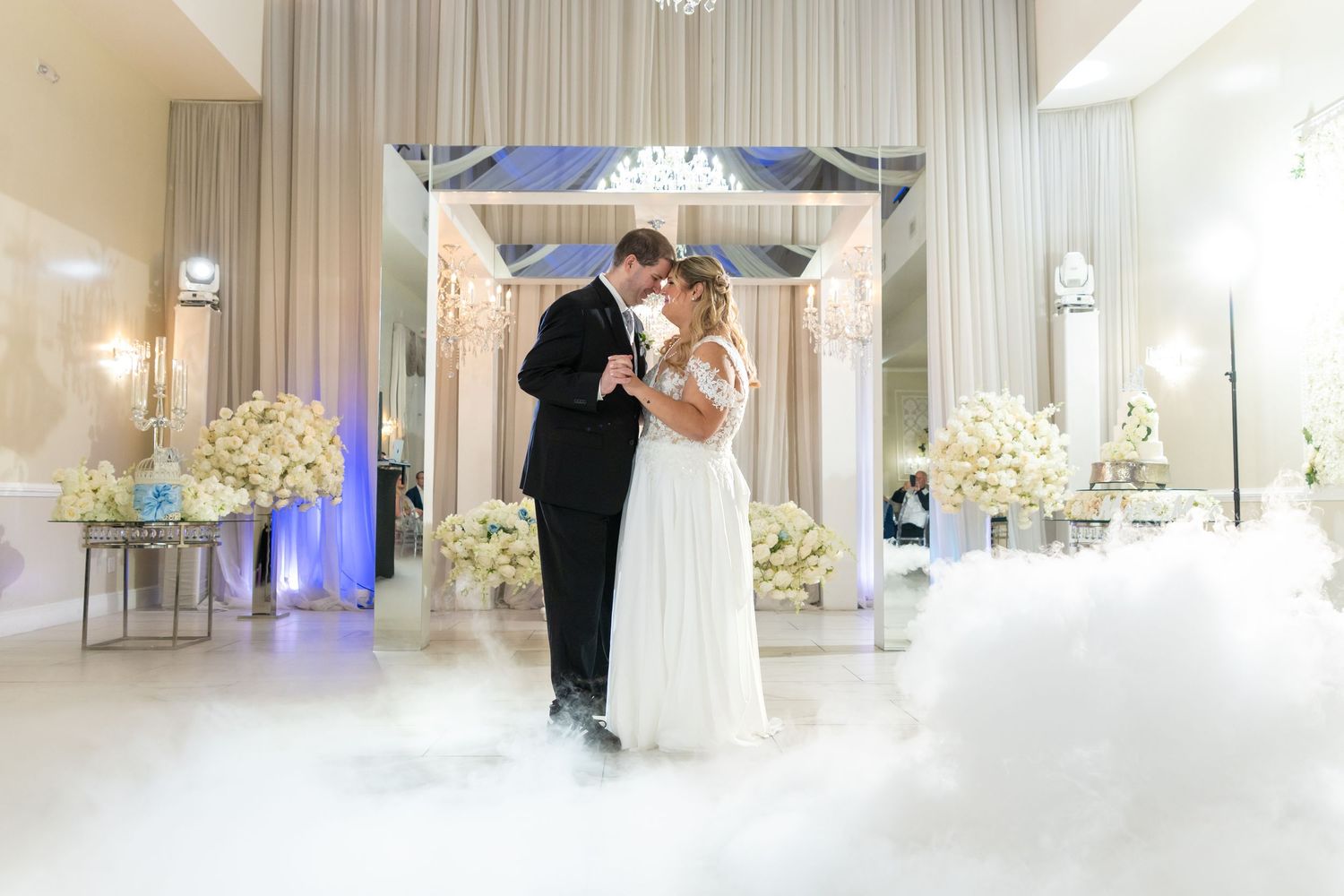 Bride and groom share first dance surrounded by white floral arrangements and dreamy fog on elegant wedding reception floor.