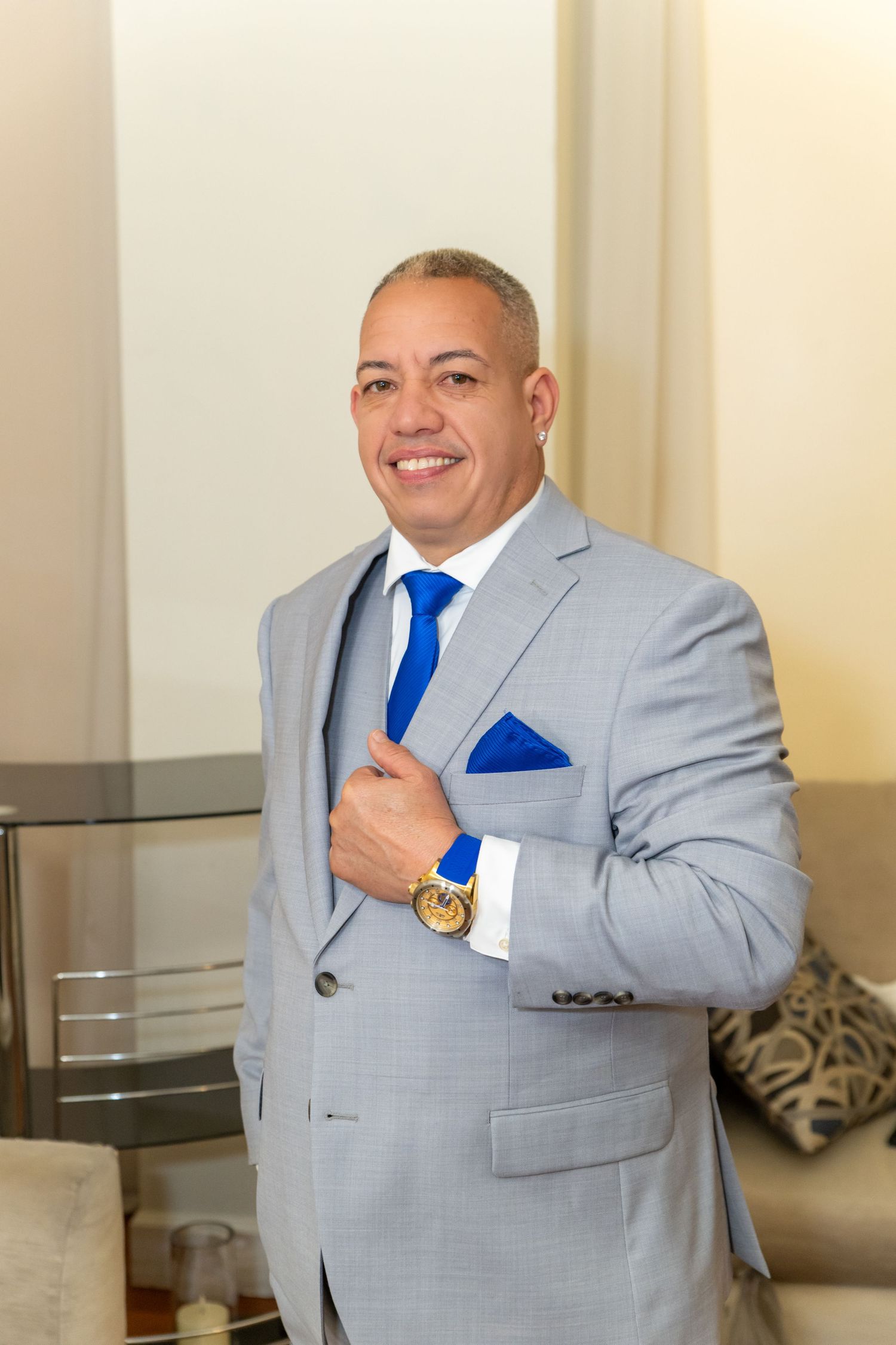 Smiling man in gray suit with blue tie and pocket square, wearing gold watch, posing confidently indoors.
