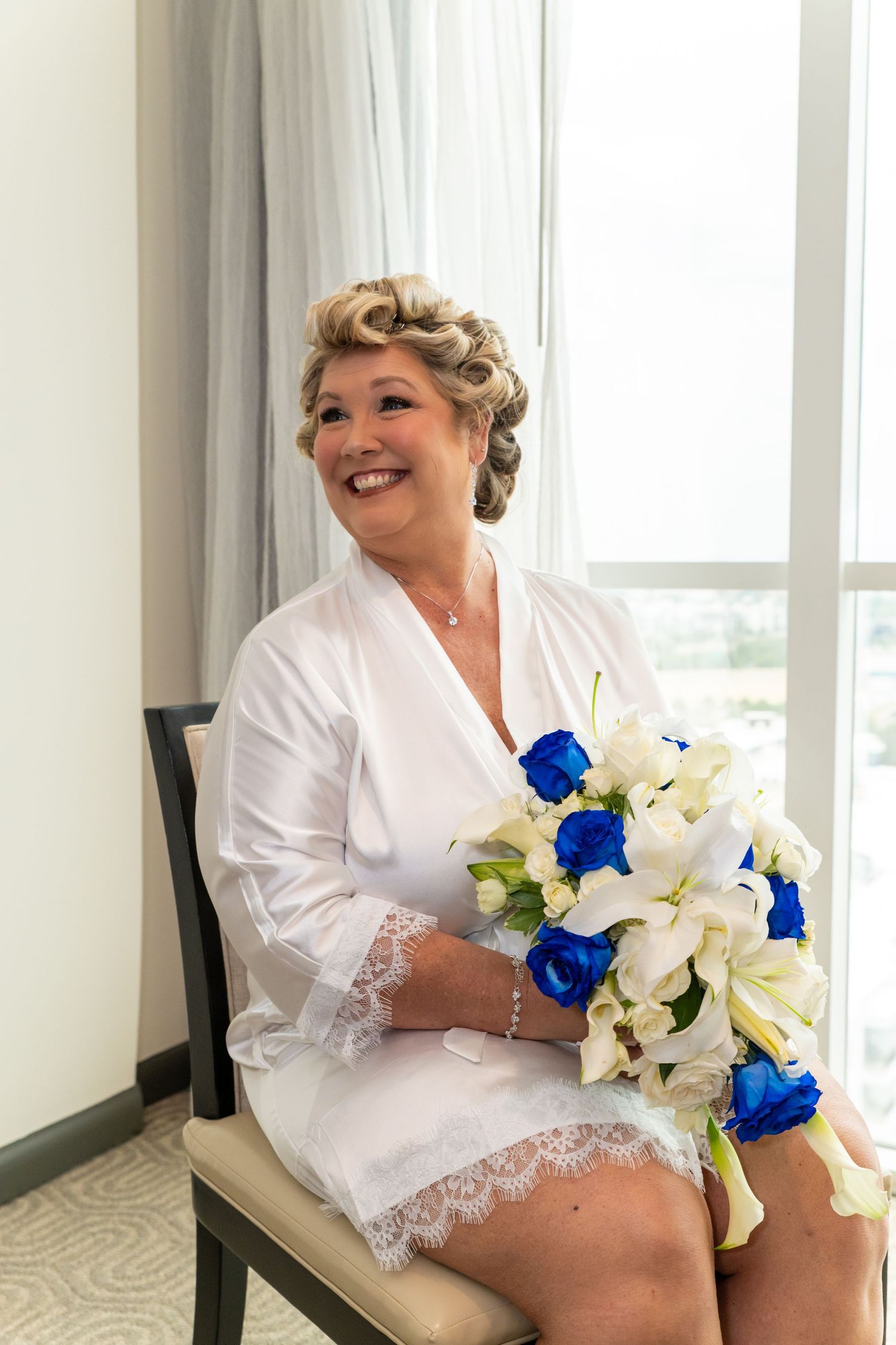 Smiling bride in white lace robe holding blue and white wedding bouquet seated by window.