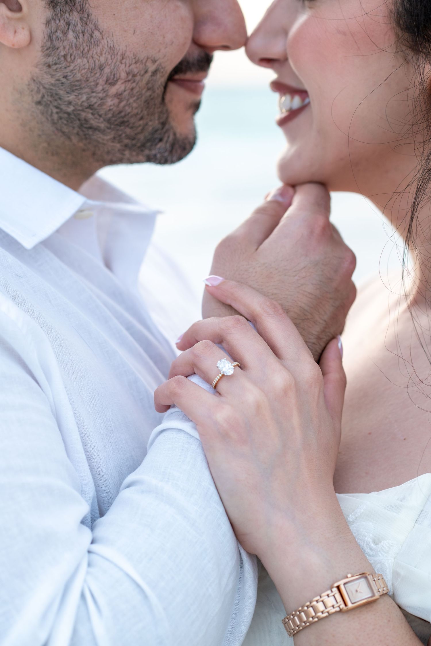 Couple nearly kissing, woman showing diamond engagement ring and rose gold watch, both dressed in white.