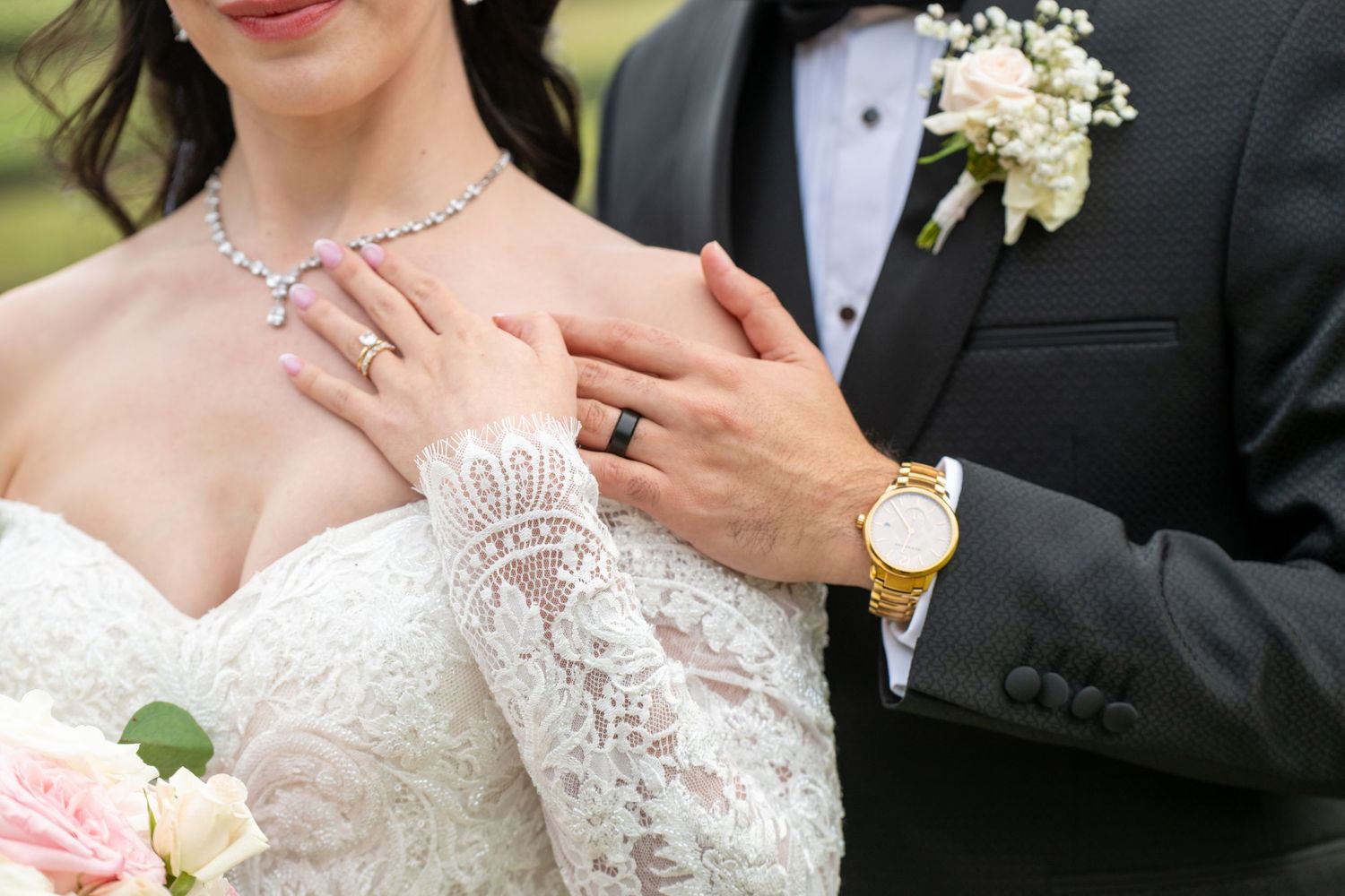 Bride in lace gown with diamond necklace and ring poses with groom in gray suit at wedding ceremony.