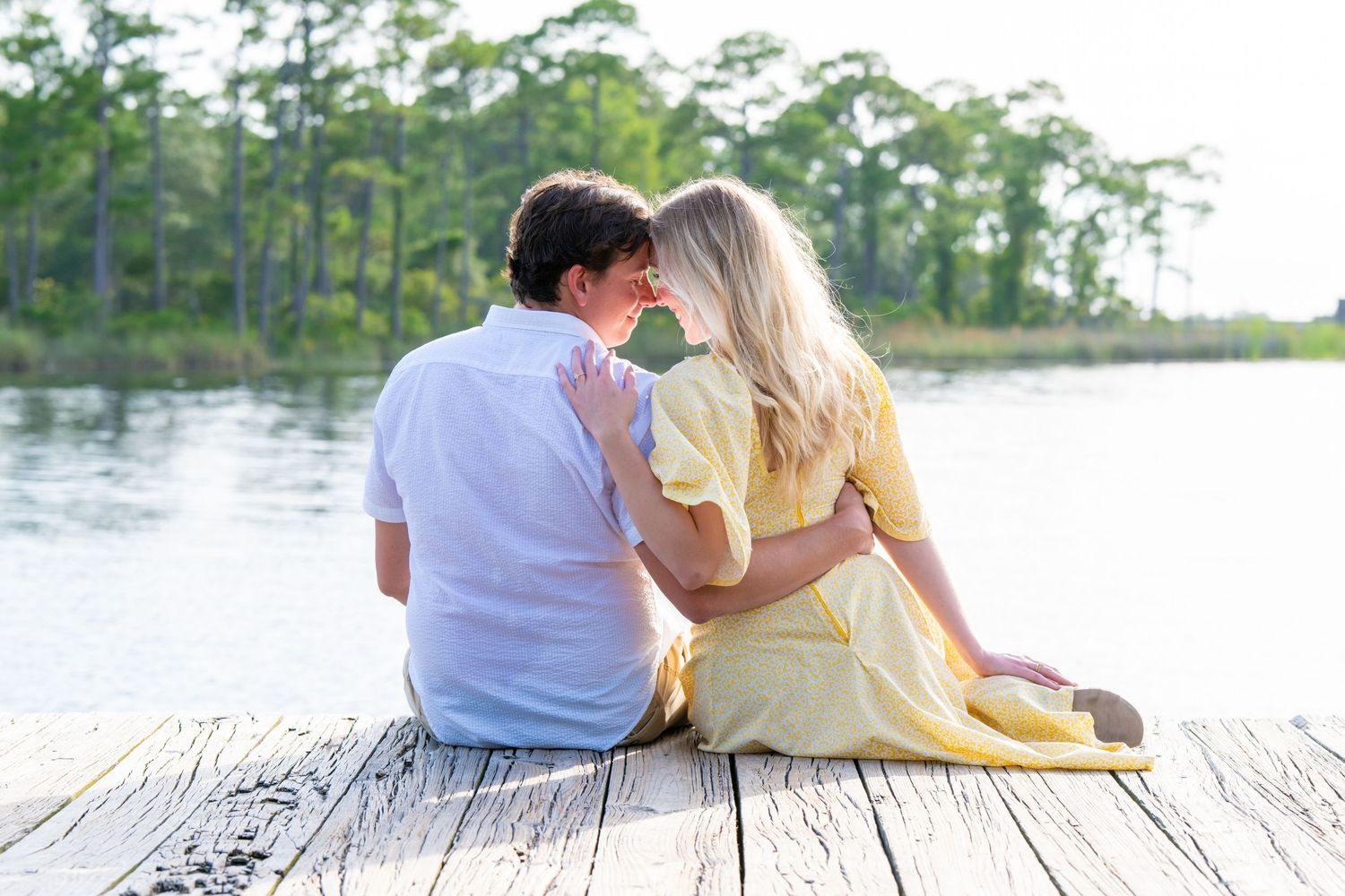 Couple sitting on a wooden dock by a lake, sharing a romantic moment surrounded by lush green trees.