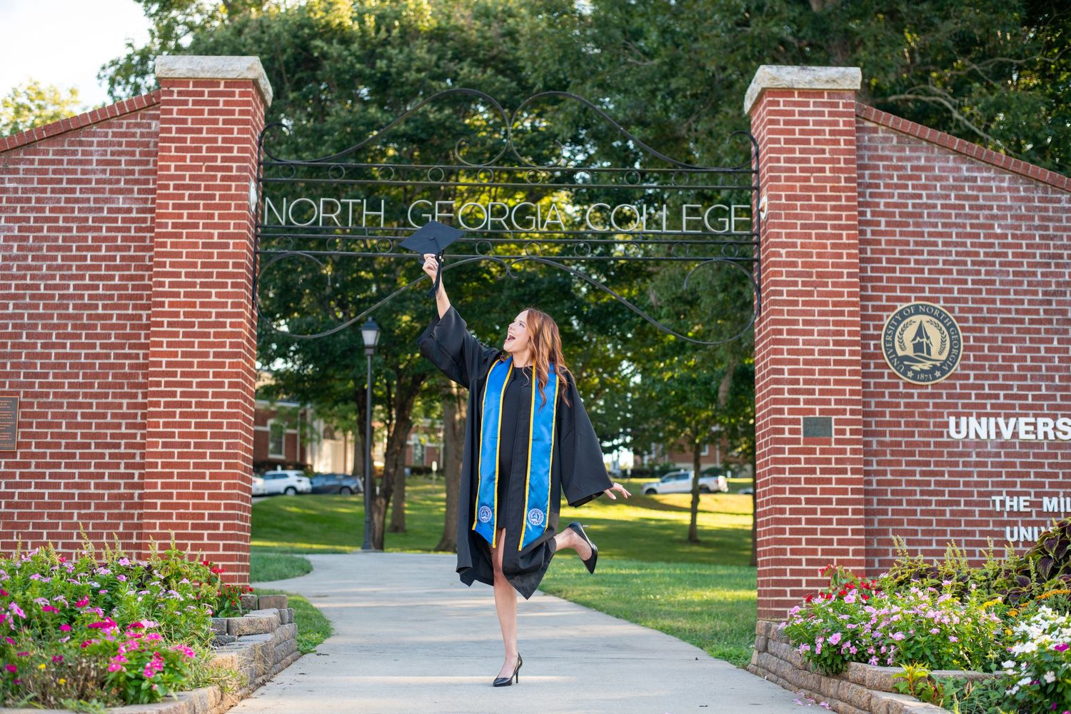 Graduate in cap and gown celebrating at North Georgia College University entrance gate on a sunny day.
