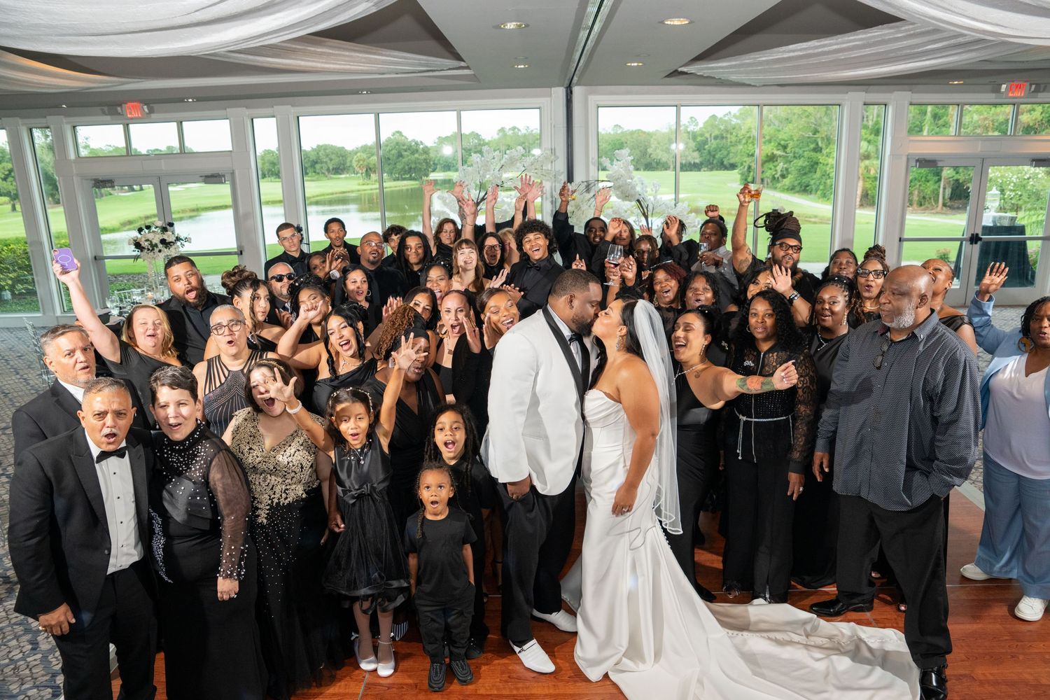 A joyful wedding group photo with bride in white gown and groom in white suit surrounded by guests in elegant black attire.