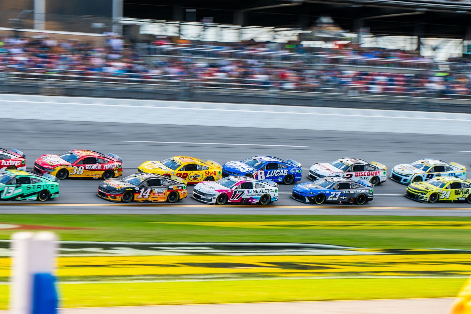 Large pack of colorful NASCAR stock cars racing side by side on superspeedway.
