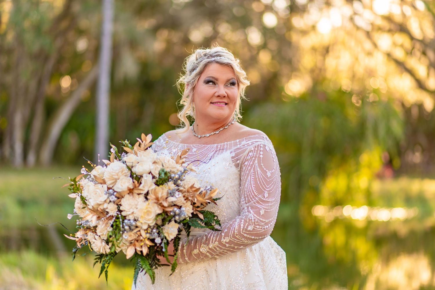 Bride in lace long-sleeve wedding gown holding blush floral bouquet, smiling outdoors at golden hour.