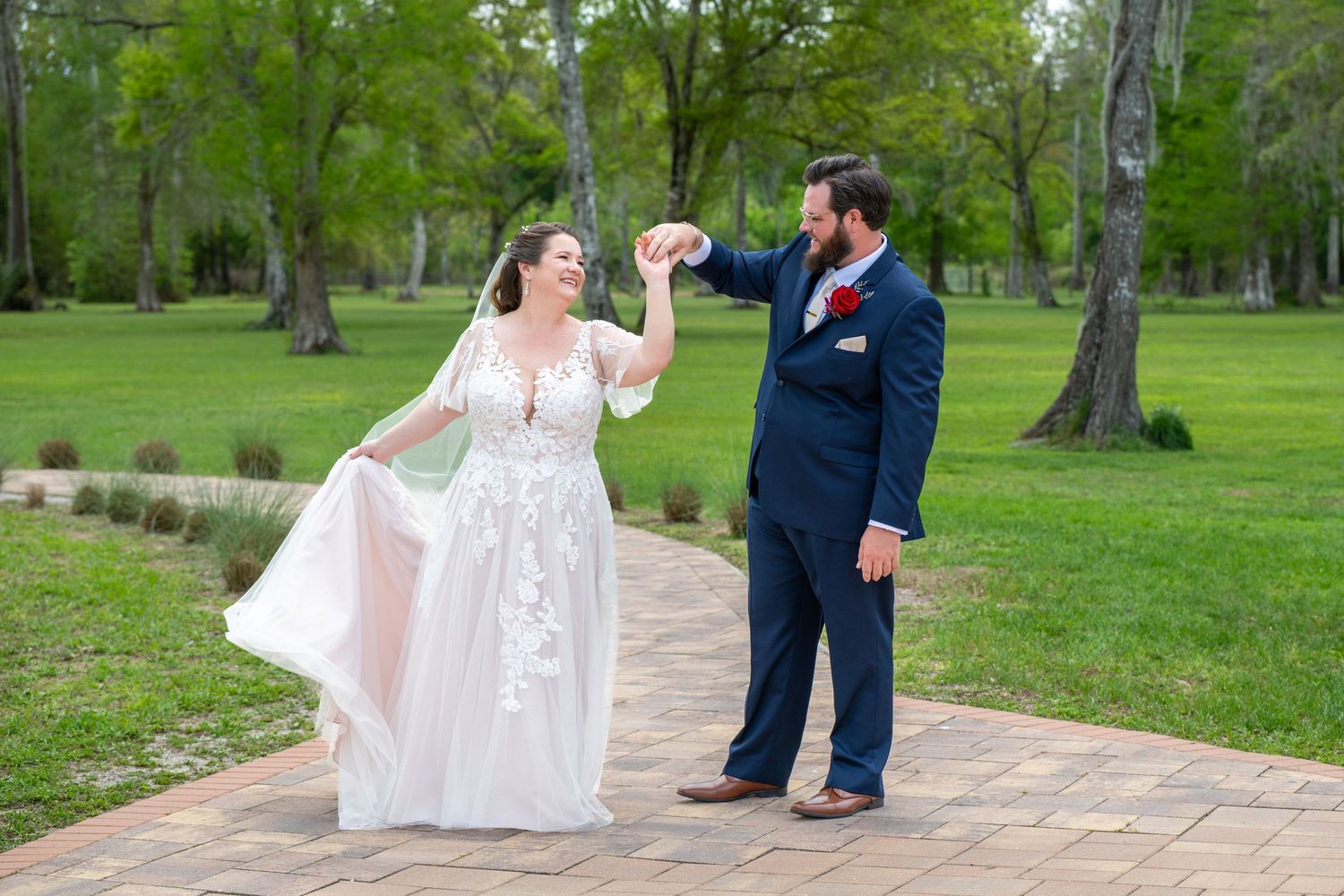 Groom twirls bride in a flowy lace wedding gown on a brick path surrounded by lush green grass.
