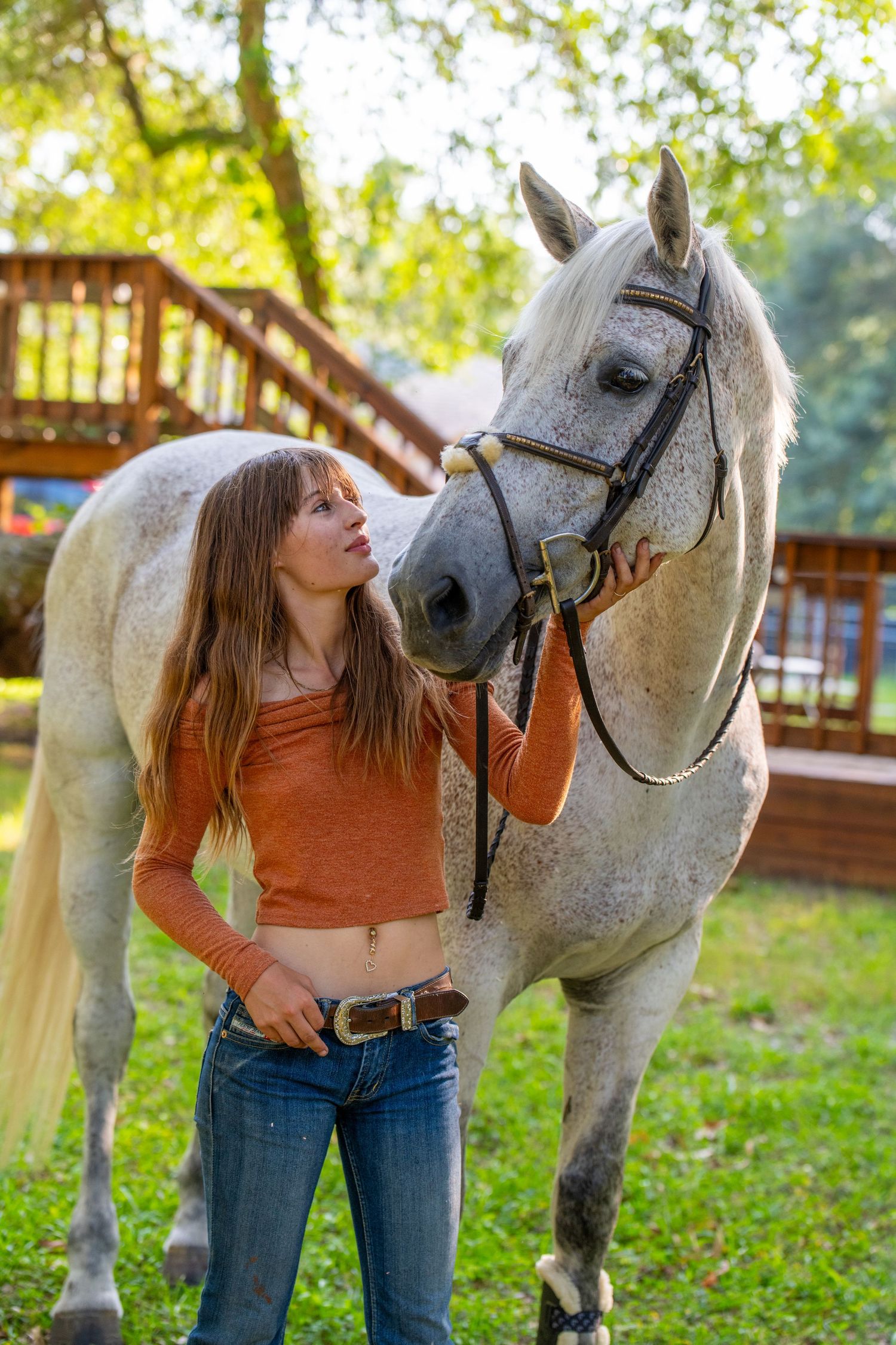 Young woman in orange top standing close to a white dapple grey horse in backyard.