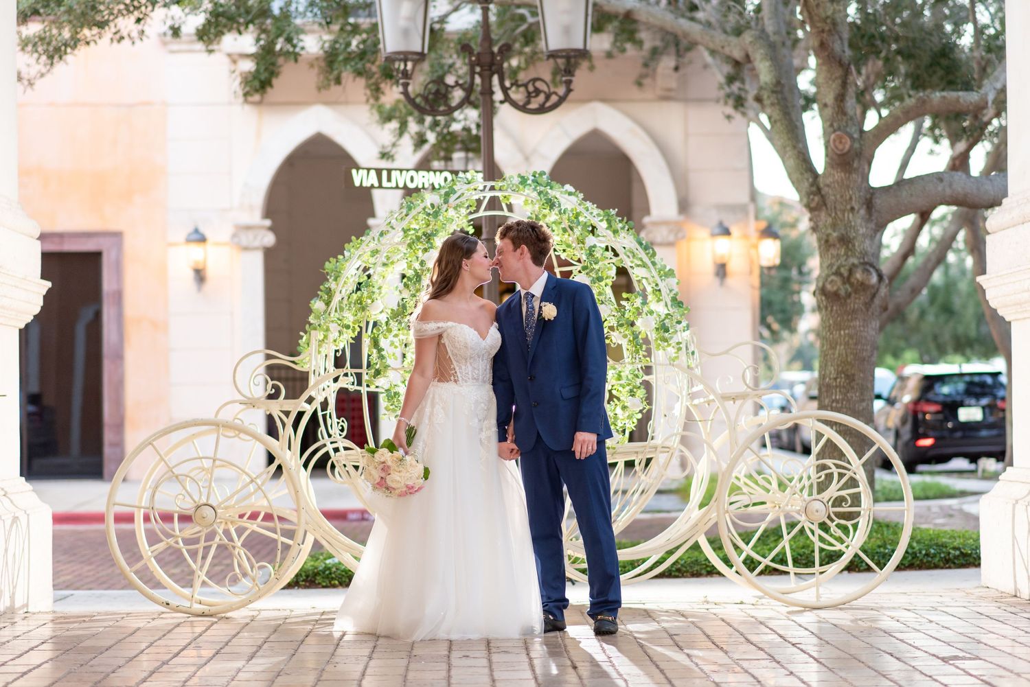 Bride and groom kissing in front of a fairy tale Cinderella carriage arch with greenery at outdoor wedding venue.