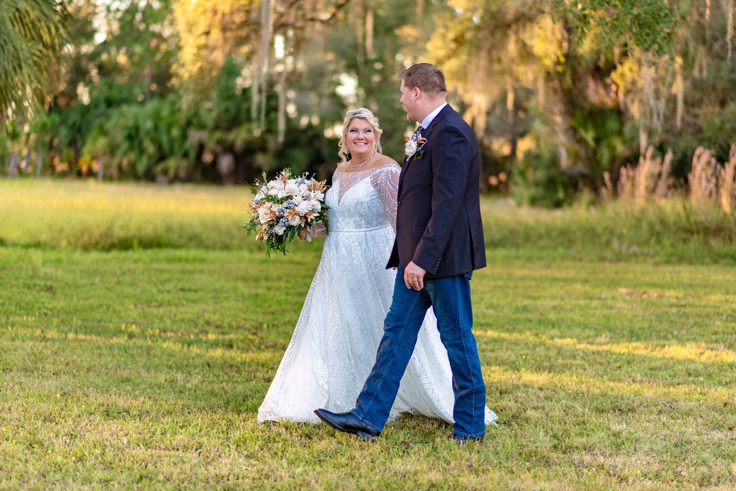 Smiling bride in white gown walks with groom in navy suit across a sunlit green field at sunset.