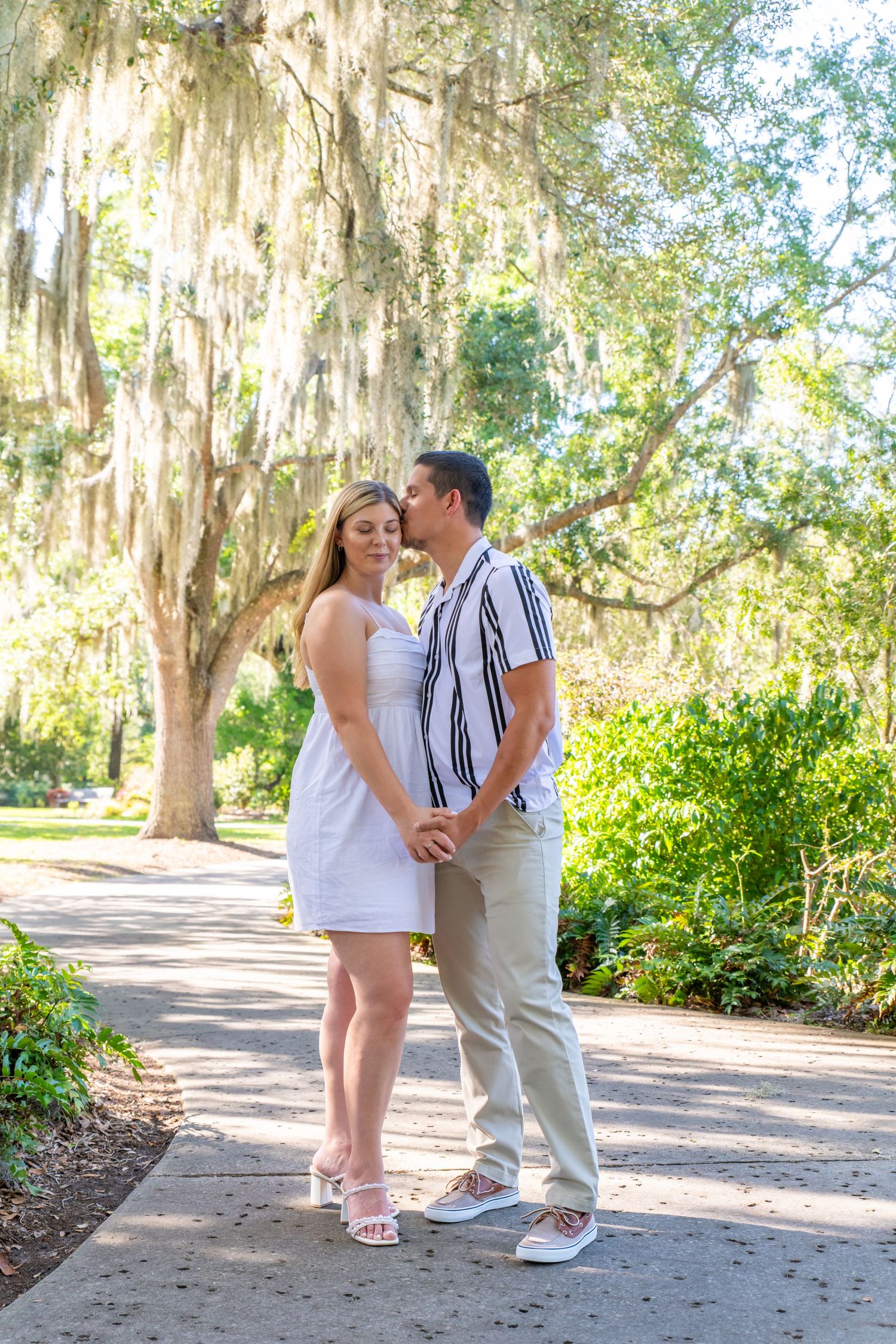 Couple holds hands and gazes lovingly at each other under Spanish moss-draped oak trees in park.