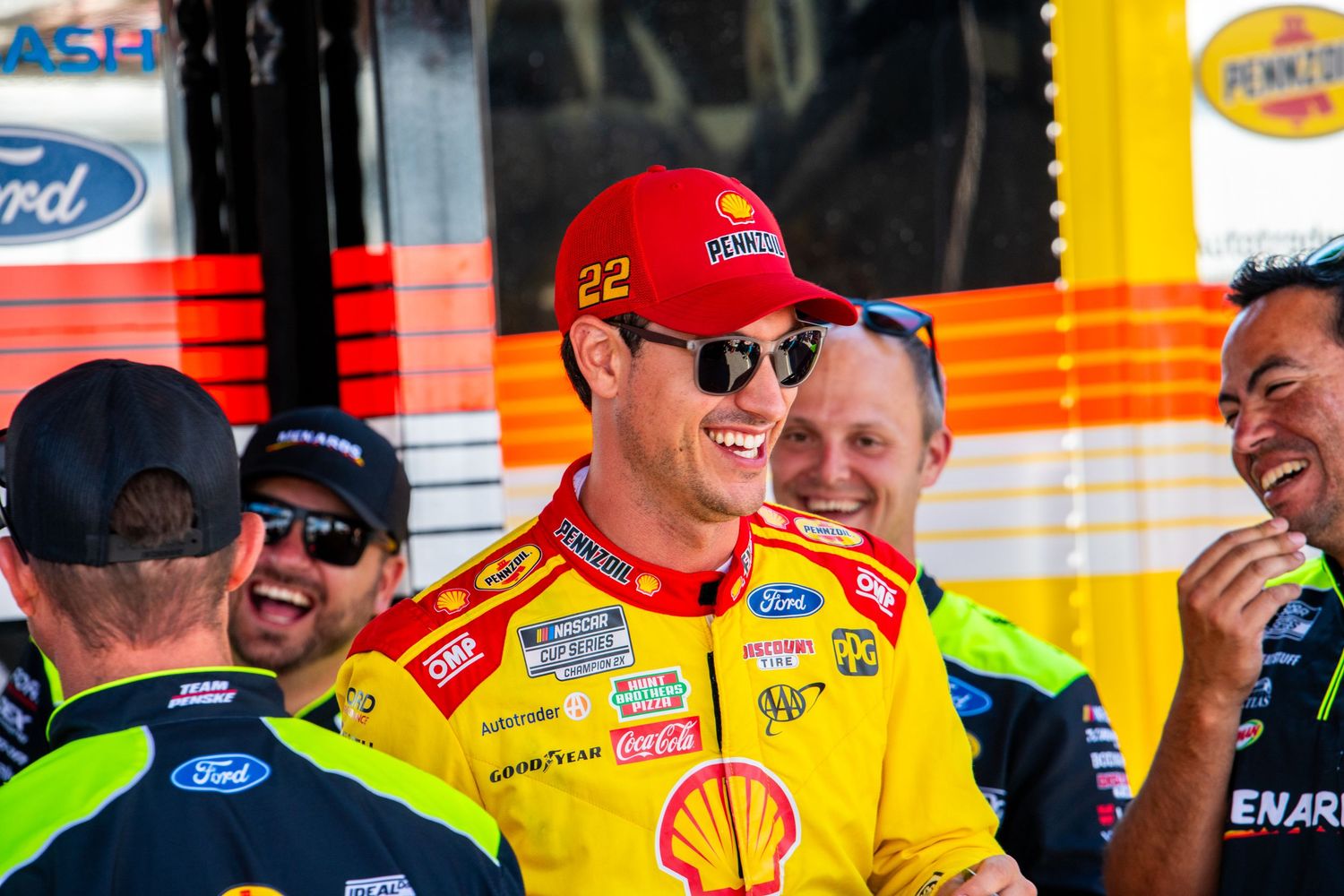 NASCAR driver wearing number 22 Shell Pennzoil cap smiling with team members in pit area. Joey Logano