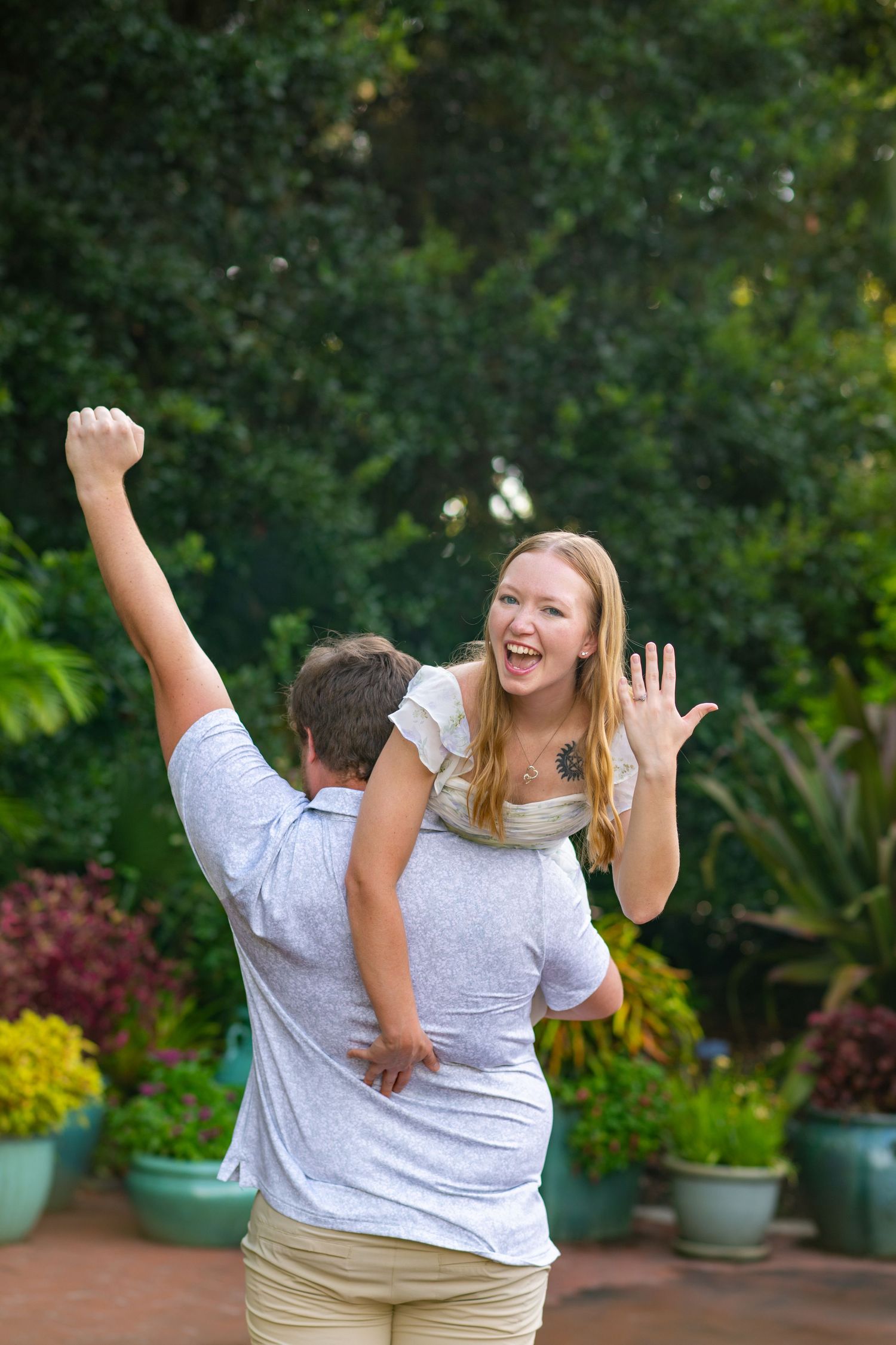 Excited woman shows off engagement ring while being lifted by her partner in a garden.