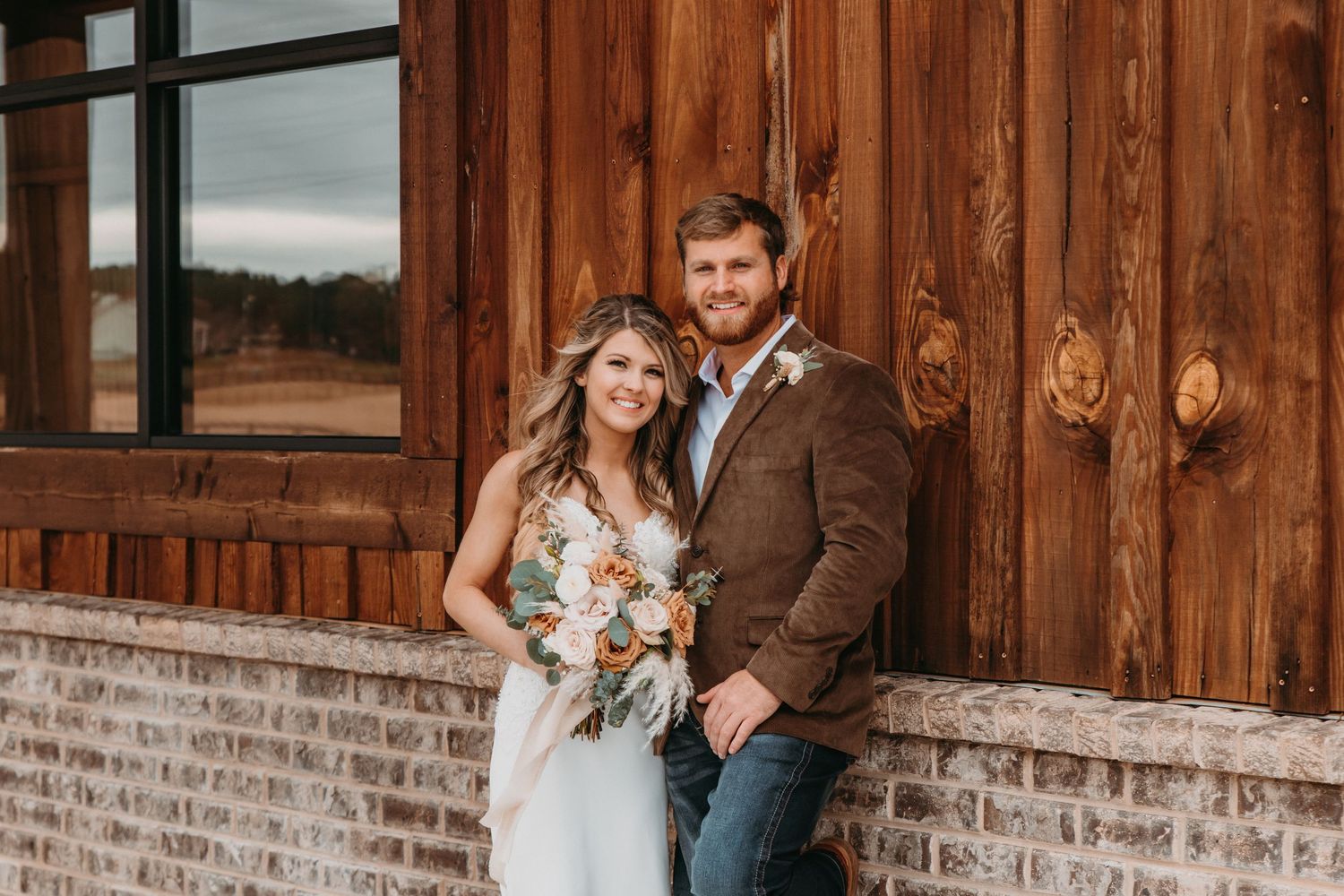 A bride and groom pose together in front of a rustic wooden barn, she holds a boho floral bouquet.