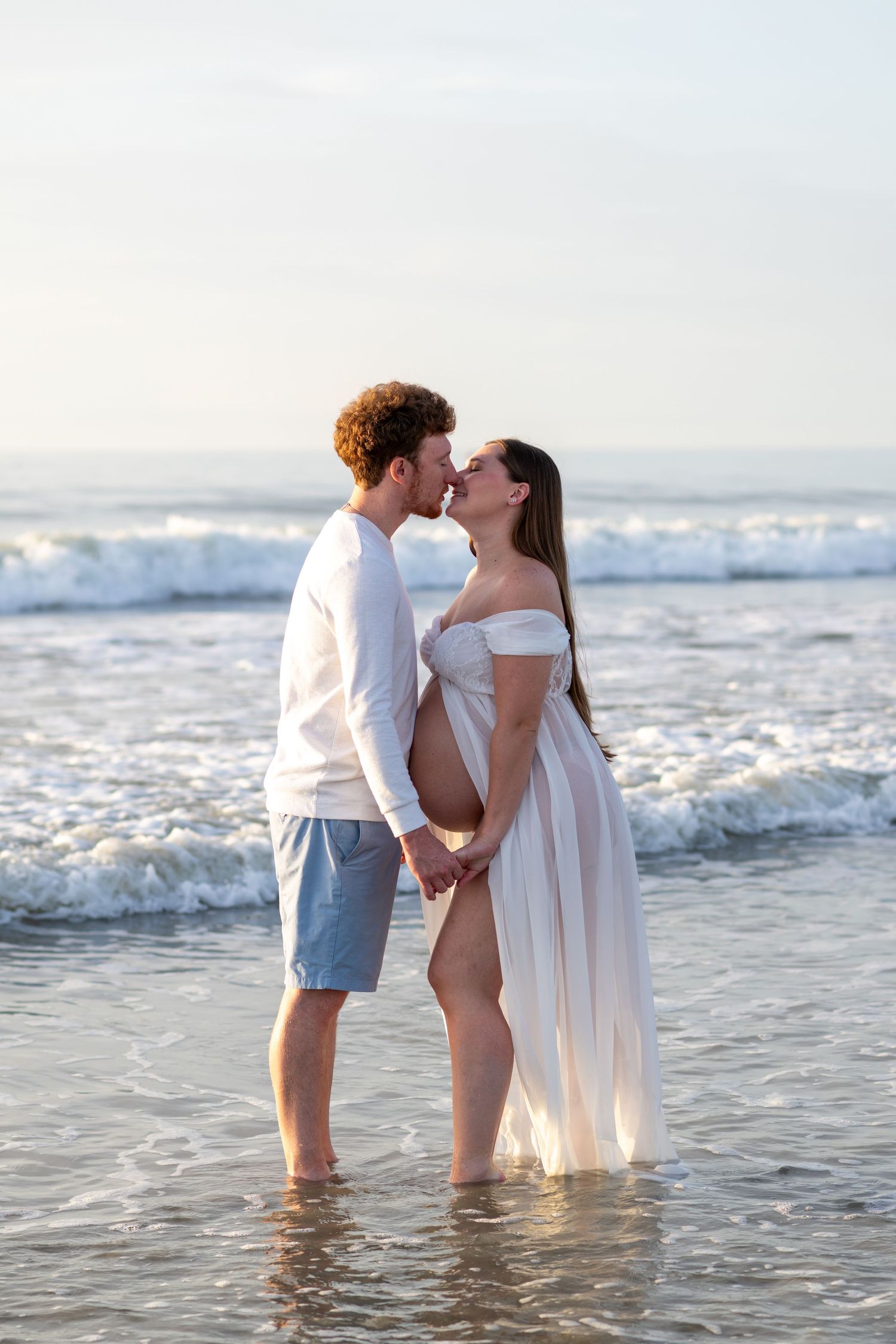 Pregnant couple kissing while standing in ocean waves at sunset, woman wearing a flowing white dress.