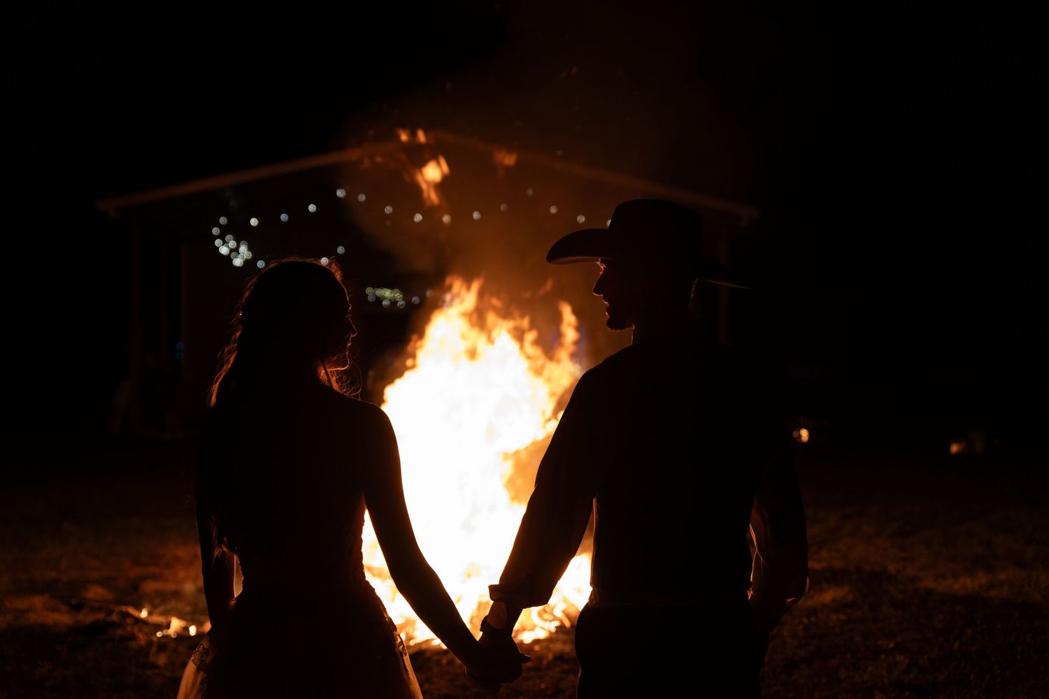 Newlyweds holding hands silhouetted against a large bonfire at night.