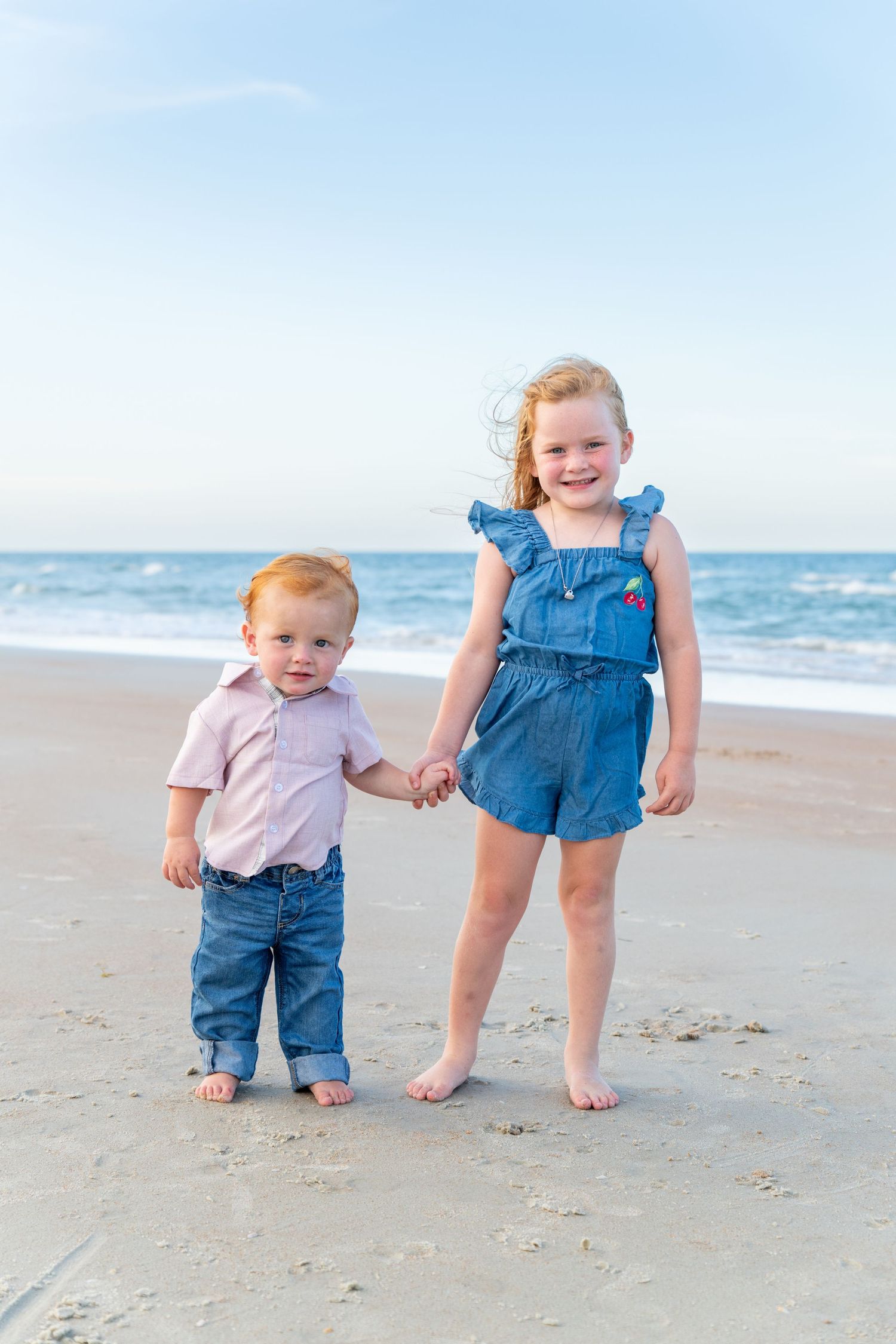 Two young siblings holding hands on a sandy beach, girl in denim romper and toddler boy in pink shirt and jeans.