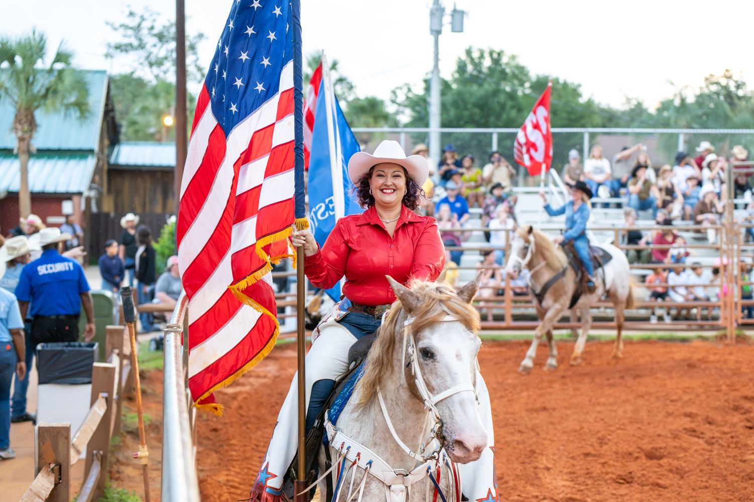 Woman in red shirt rides white horse carrying American flag at outdoor rodeo arena with spectators in background.