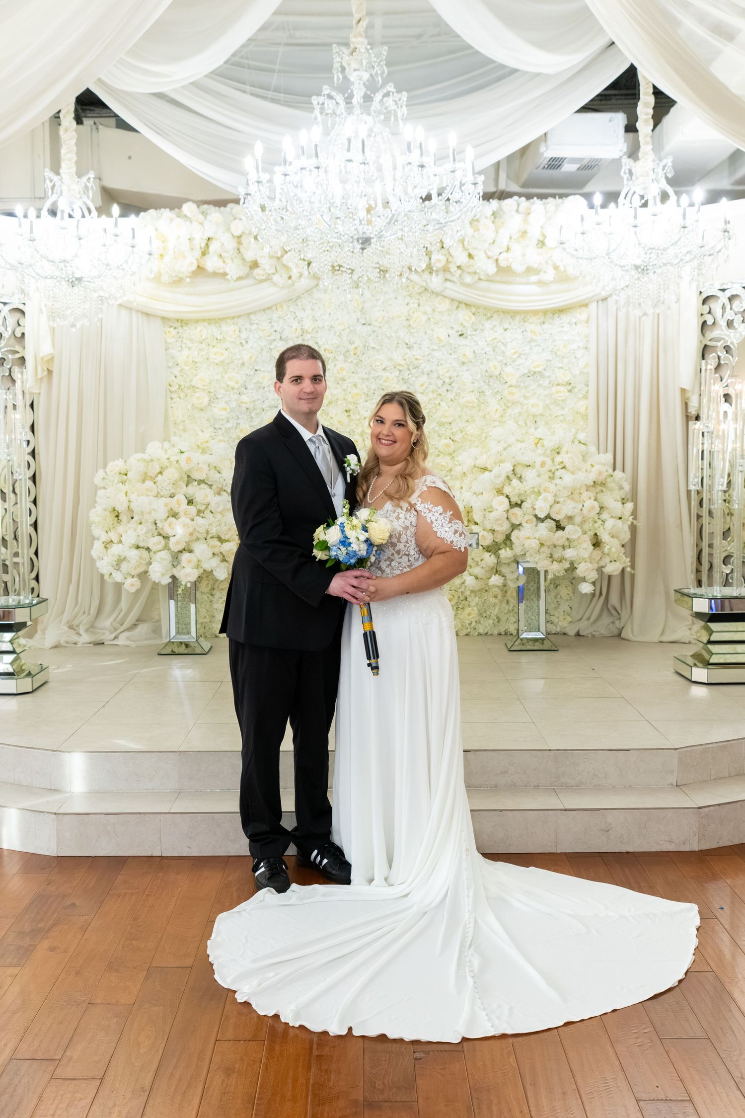 Bride and groom posing in front of white floral backdrop with crystal chandelier in elegant wedding venue.