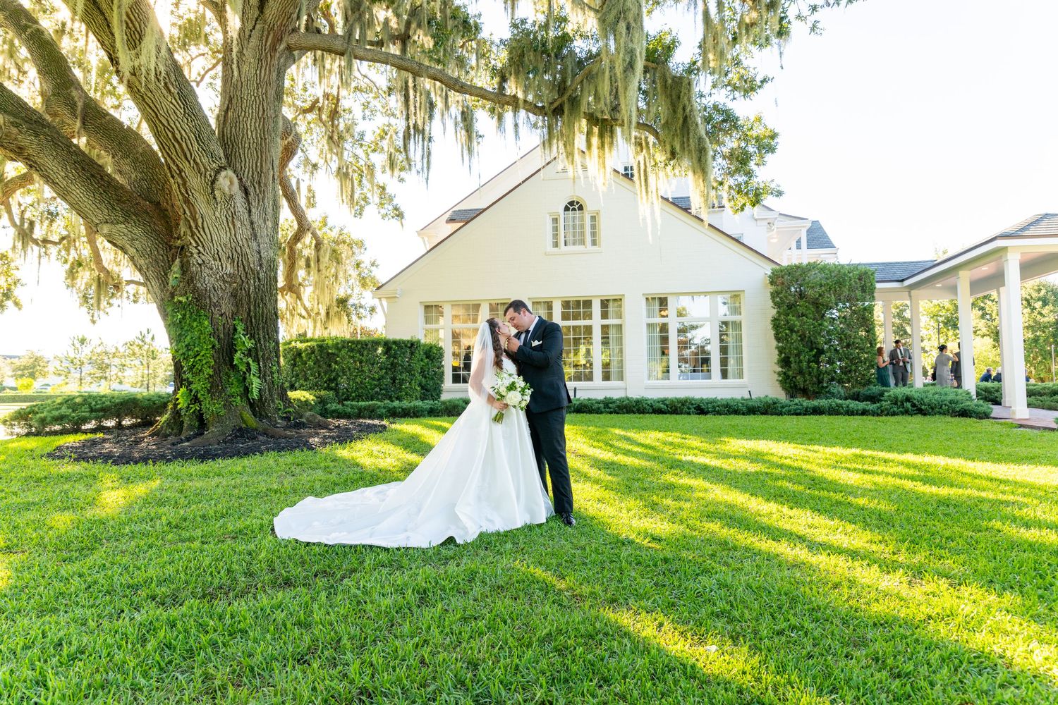 Bride and groom share a romantic kiss on lush green lawn beside a grand oak tree at an elegant wedding venue.