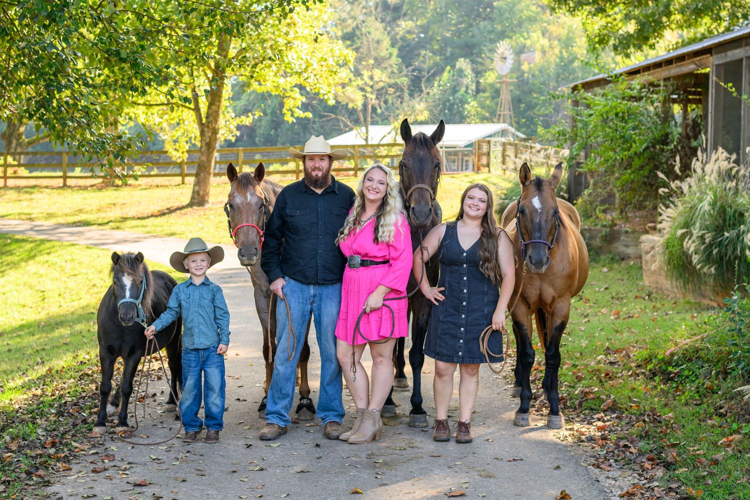 Family of four posing with three horses on a farm, wearing western attire including cowboy hats and boots.