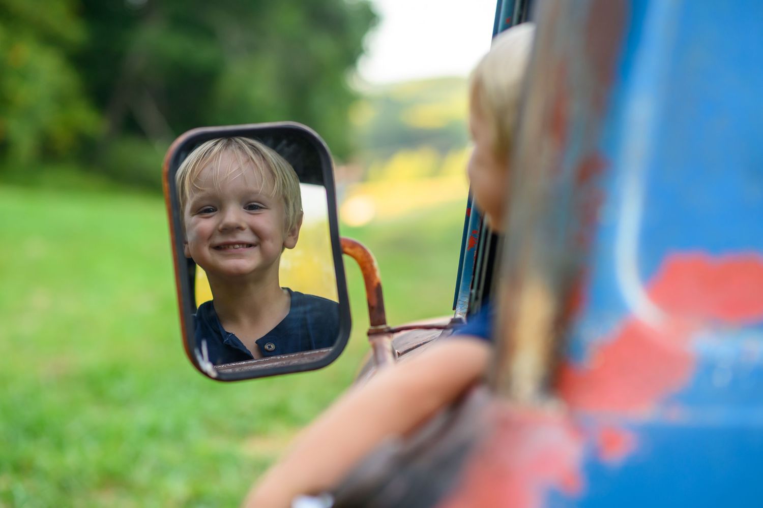 Smiling blond boy reflected in a vintage truck side mirror with green countryside in the background.
