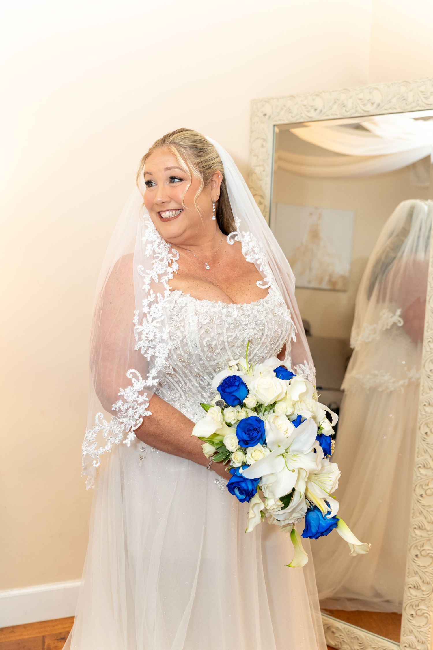 Smiling bride in white lace gown and veil holds blue and white bouquet, standing near ornate mirror.