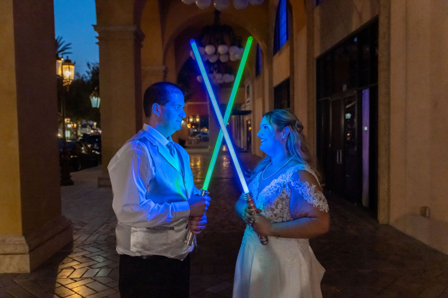Bride and groom holding glowing Star Wars lightsabers in blue and green at an outdoor evening wedding celebration.