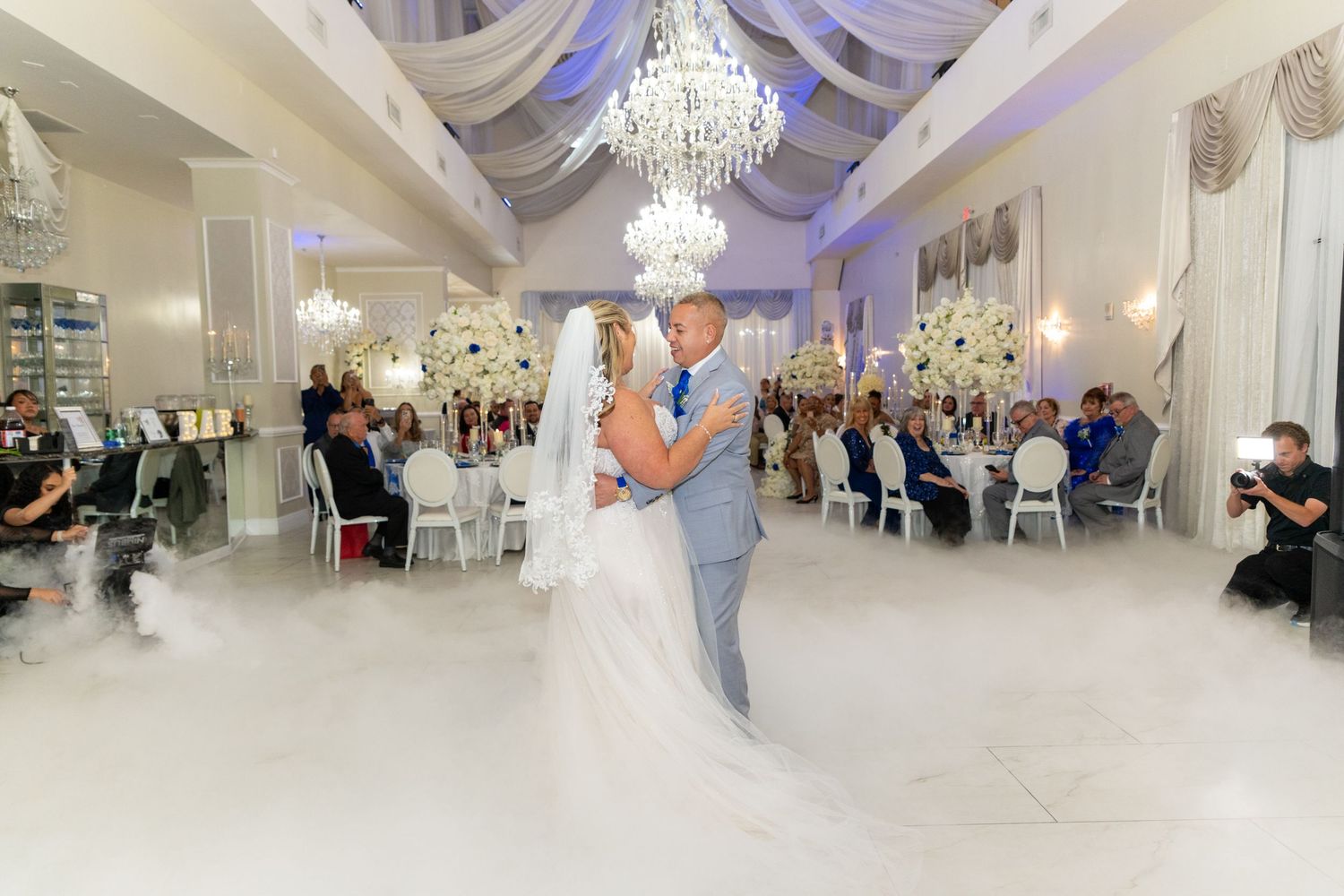Bride and groom share first dance on fog-covered floor in elegant white ballroom with crystal chandeliers and seated wedding guests.